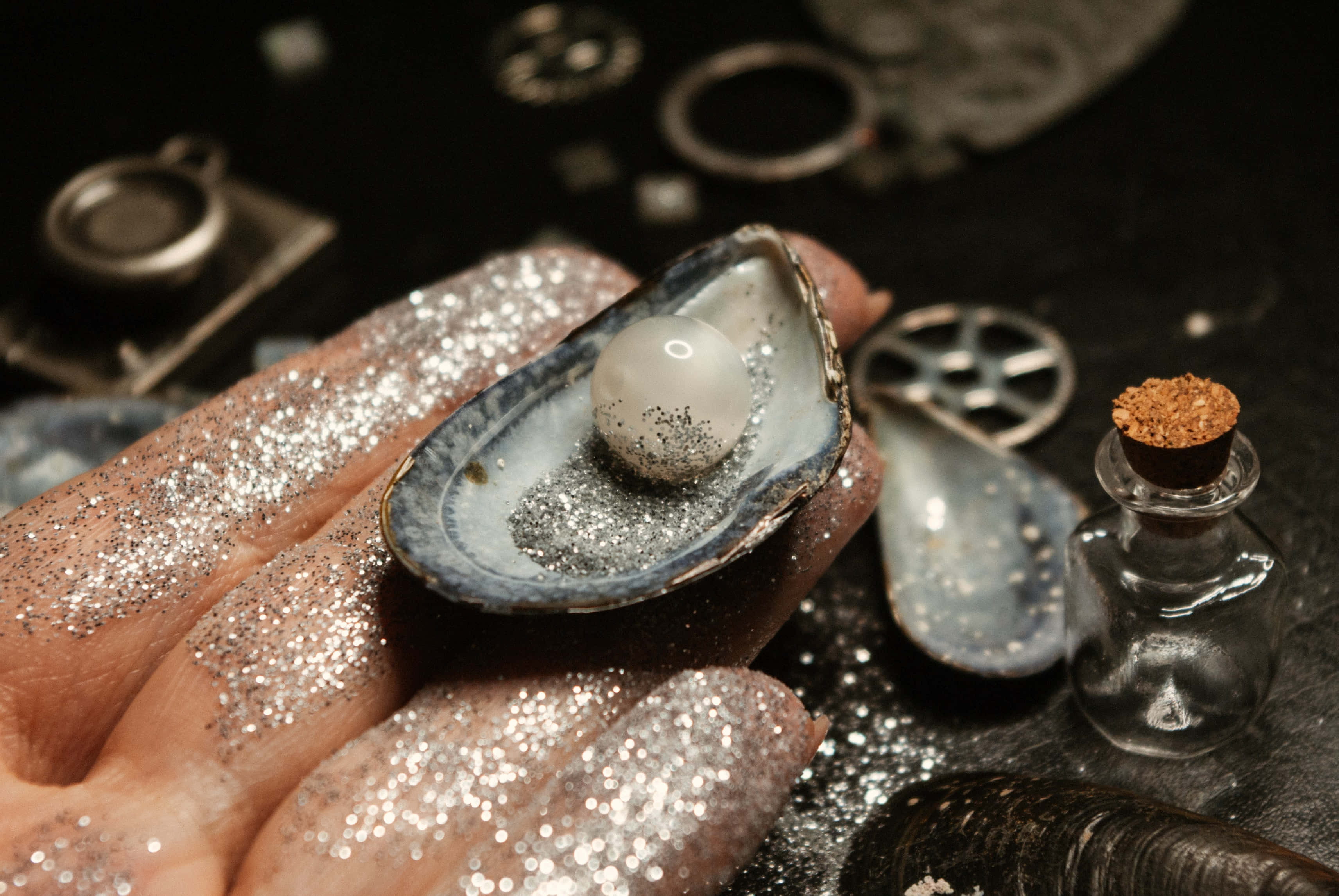 pearl 桌布 壁紙 — 一隻佈滿銀色閃粉的手，捧著一個打開的牡蠣殼，裡面有一顆發光的珍珠，背景是深色的，散落著貝殼和一個小小的軟木塞瓶子。