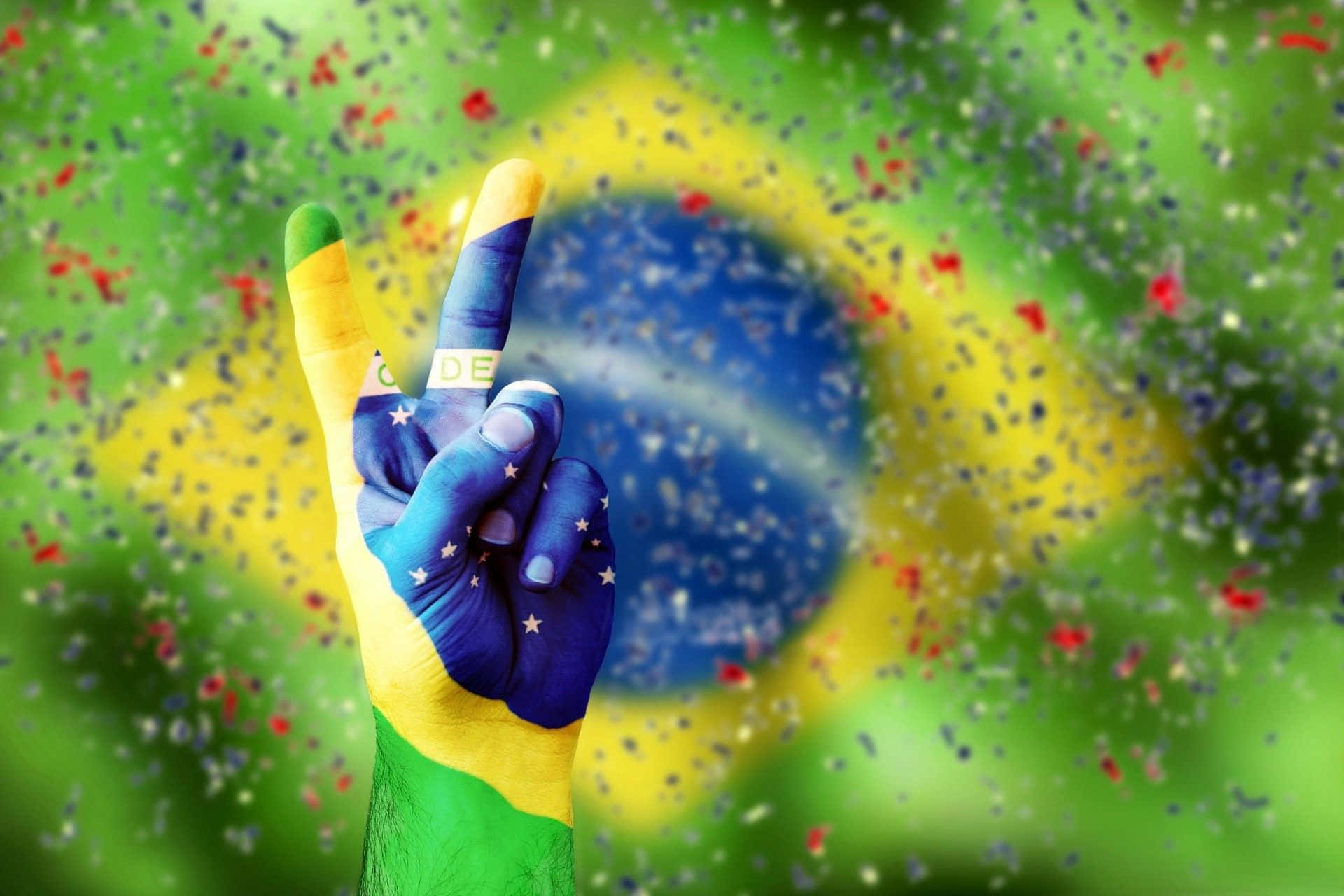 Brazil 桌布 壁紙 — 一隻塗有巴西國旗的手在模糊的旗幟背景和飄落的五彩紙屑中做出和平手勢。