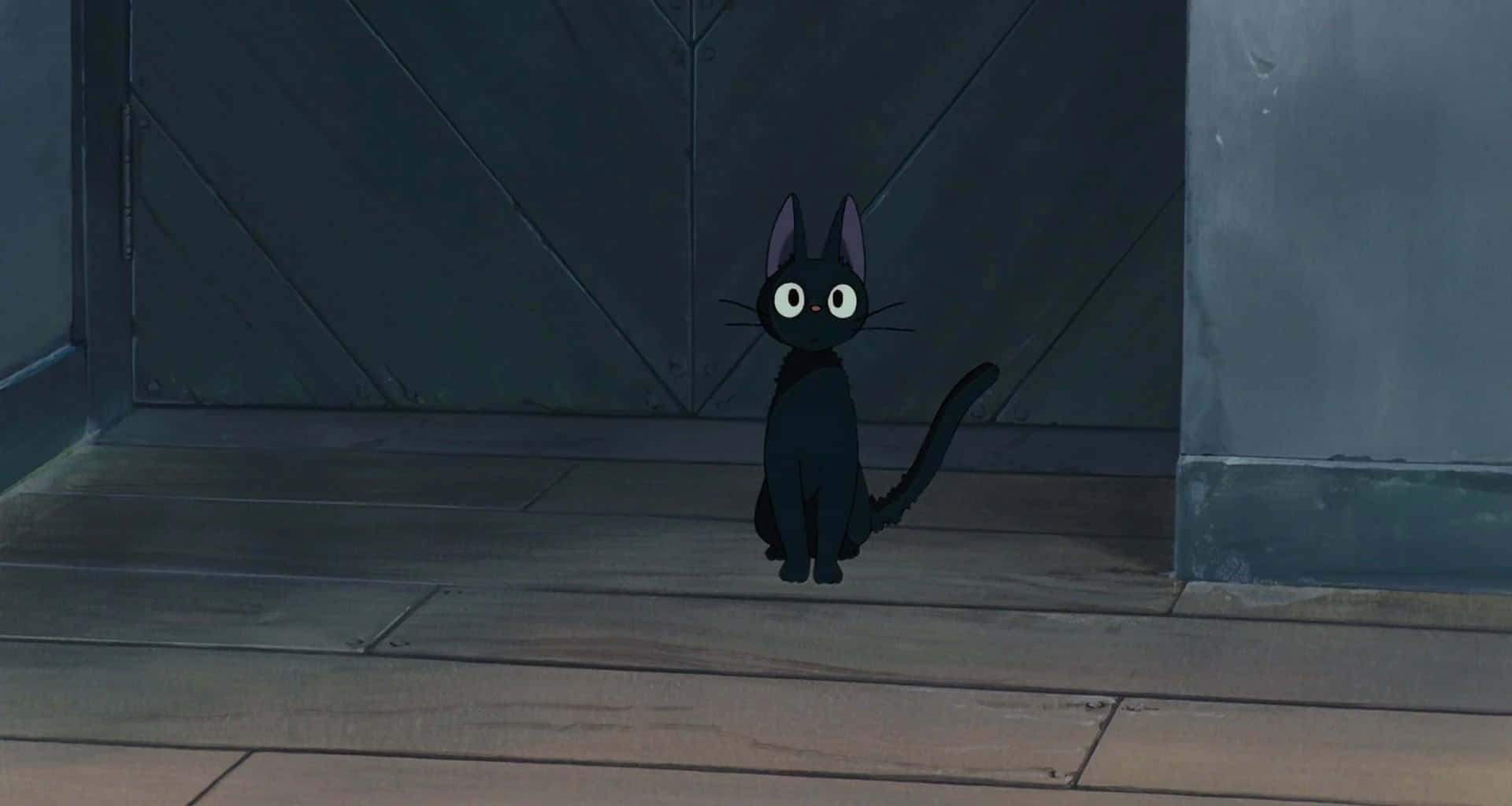 Jiji Cat 桌布 壁紙 — 一隻小黑貓睜著白色大眼睛，警覺地坐在昏暗房間的木地板上，靠近深色木門。
