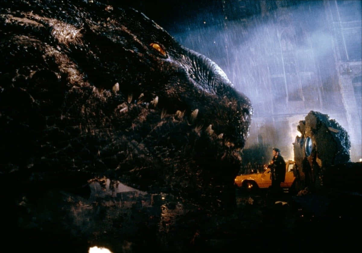 godzilla 1998 桌布 壁紙 — 一隻巨大的哥斯拉籠罩在被雨水浸透的城市街道上，一個孤獨的身影在黑暗中站在一輛黃色汽車旁邊。