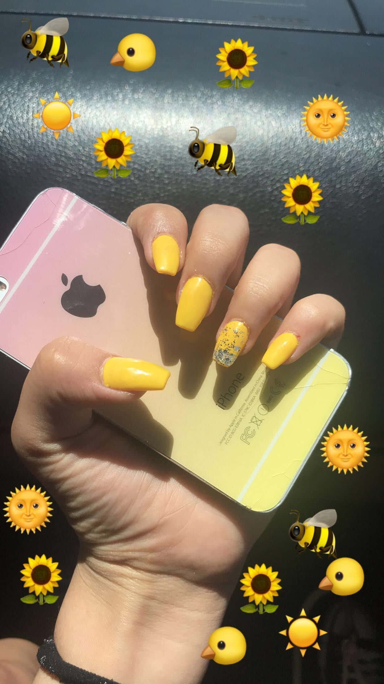 Cute Nails 桌布 壁紙 — 一隻帶有亮黃色指甲的手在深色表面上拿著一部粉紅色iPhone，周圍環繞著快樂的蜜蜂、向日葵和太陽表情符號。