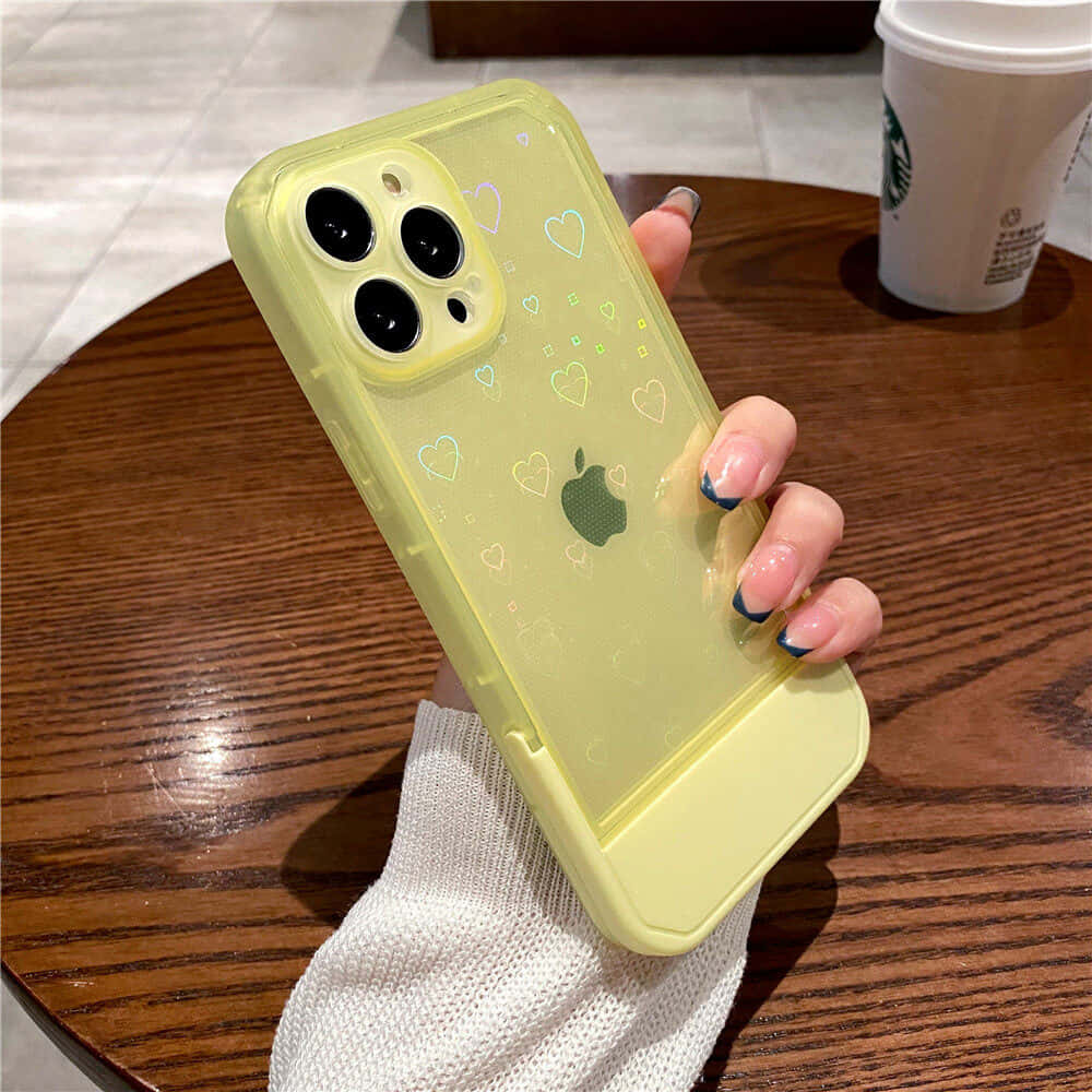 iPhone風格壁紙 桌布 壁紙 — 一隻帶有藍色指尖指甲的手，在木桌上拿著一個帶有心形貼紙的黃色手機殼，旁邊放著一杯咖啡。
