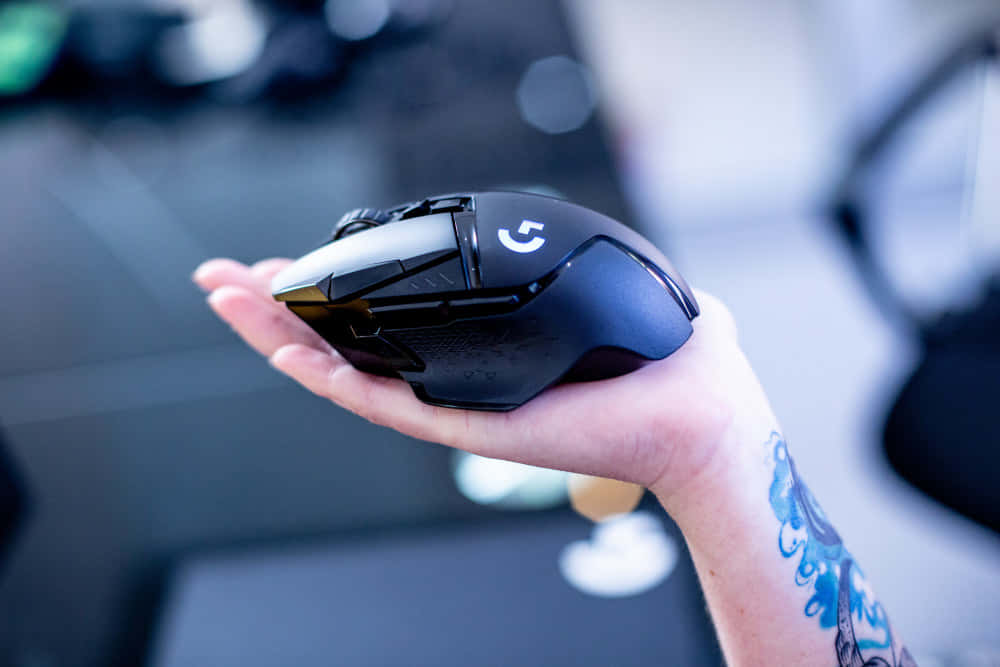 Gaming Mouse 桌布 壁紙 — 一隻帶有藍色紋身的手，在模糊的科技工作空間背景下，握著一款時尚的黑色Logitech遊戲滑鼠。