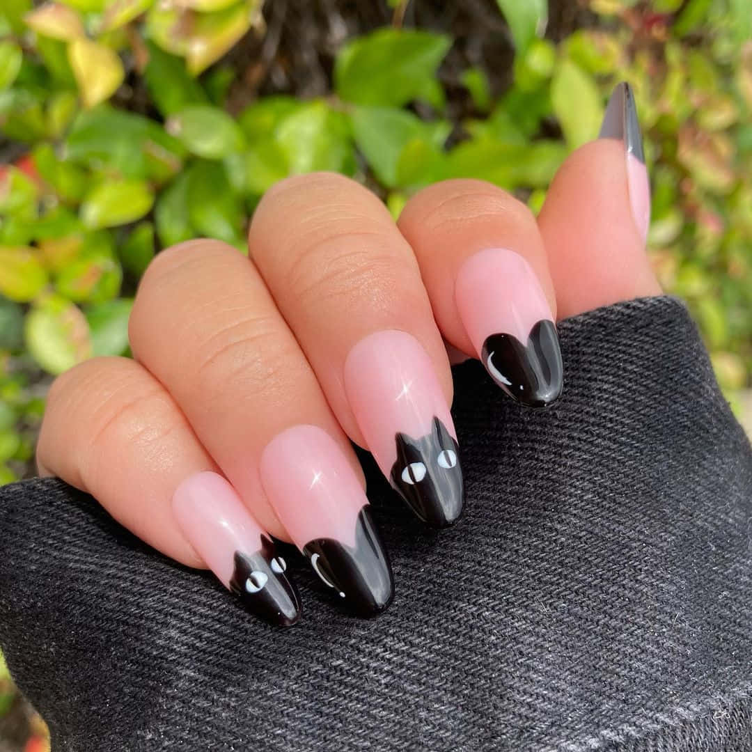 Halloween Nail Art 桌布 壁紙 — 一隻帶有長尖指甲、粉紅色指尖上有黑色貓臉的手，放在深色牛仔布上，背景是綠色樹葉。