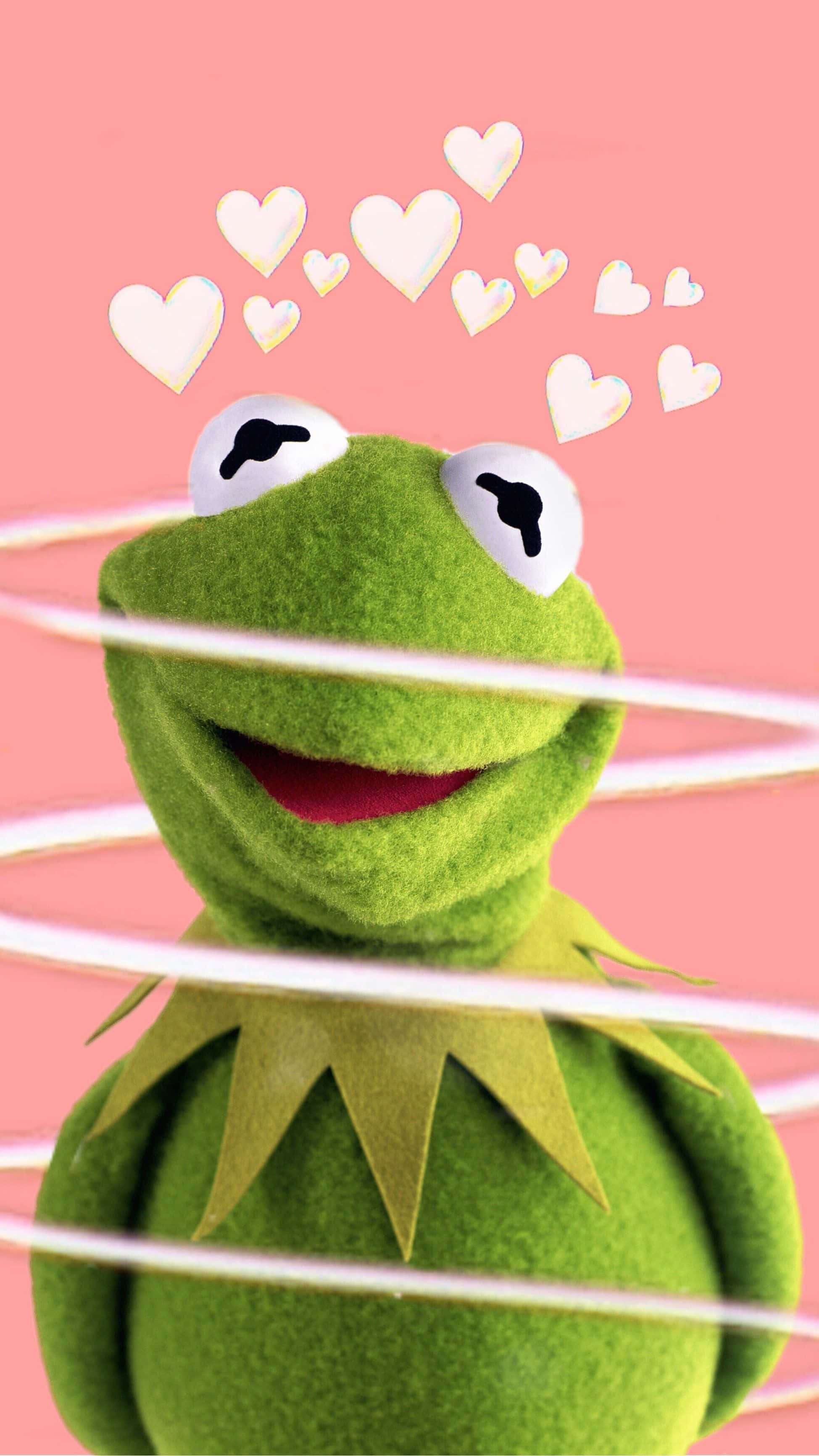 Aesthetic Frog 桌布 壁紙 — 一隻快樂的綠色青蛙木偶，頭上有心形漂浮在柔和的粉紅色背景上。
