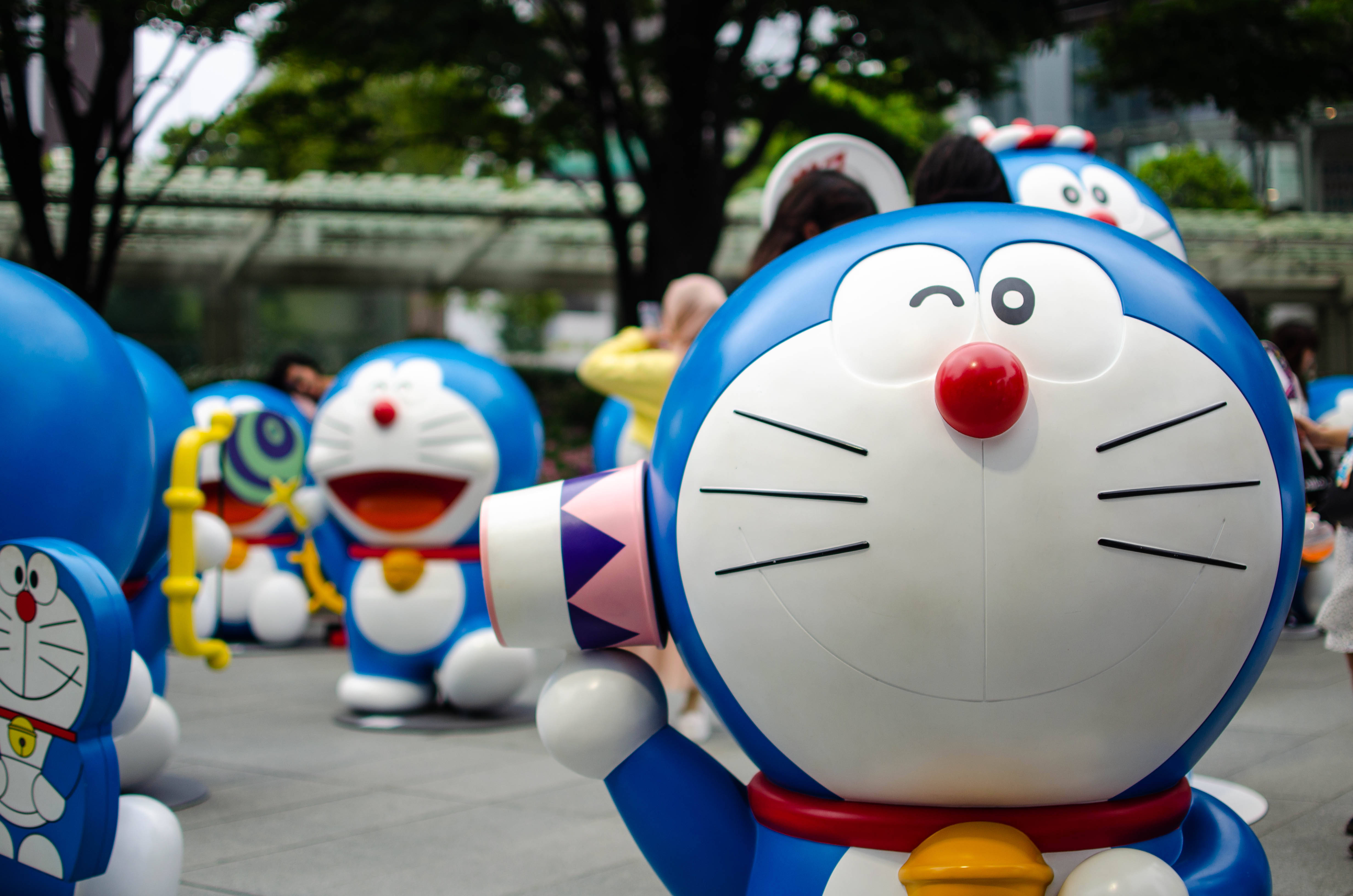 Doraemon 桌布 壁紙 — 一隻快樂的藍色機械貓，紅鼻子白臉，在陽光明媚的戶外廣場上，從一群相同的多啦A夢雕像中揮手。