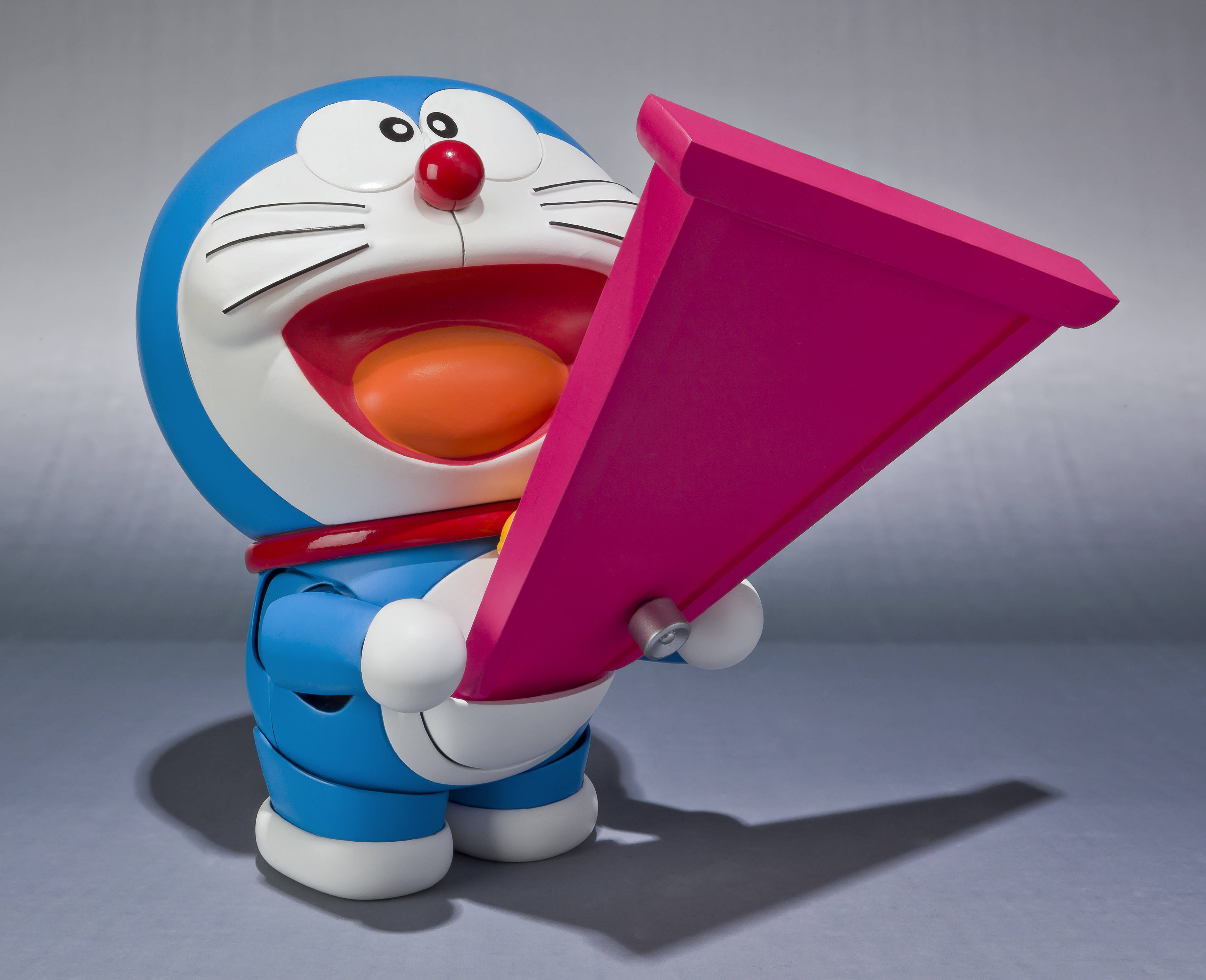 Doraemon 桌布 壁紙 — 一隻快樂的藍色機械貓，紅鼻子白臉，在柔和的灰色工作室背景前拿著一個亮粉紅色的三角形物體。