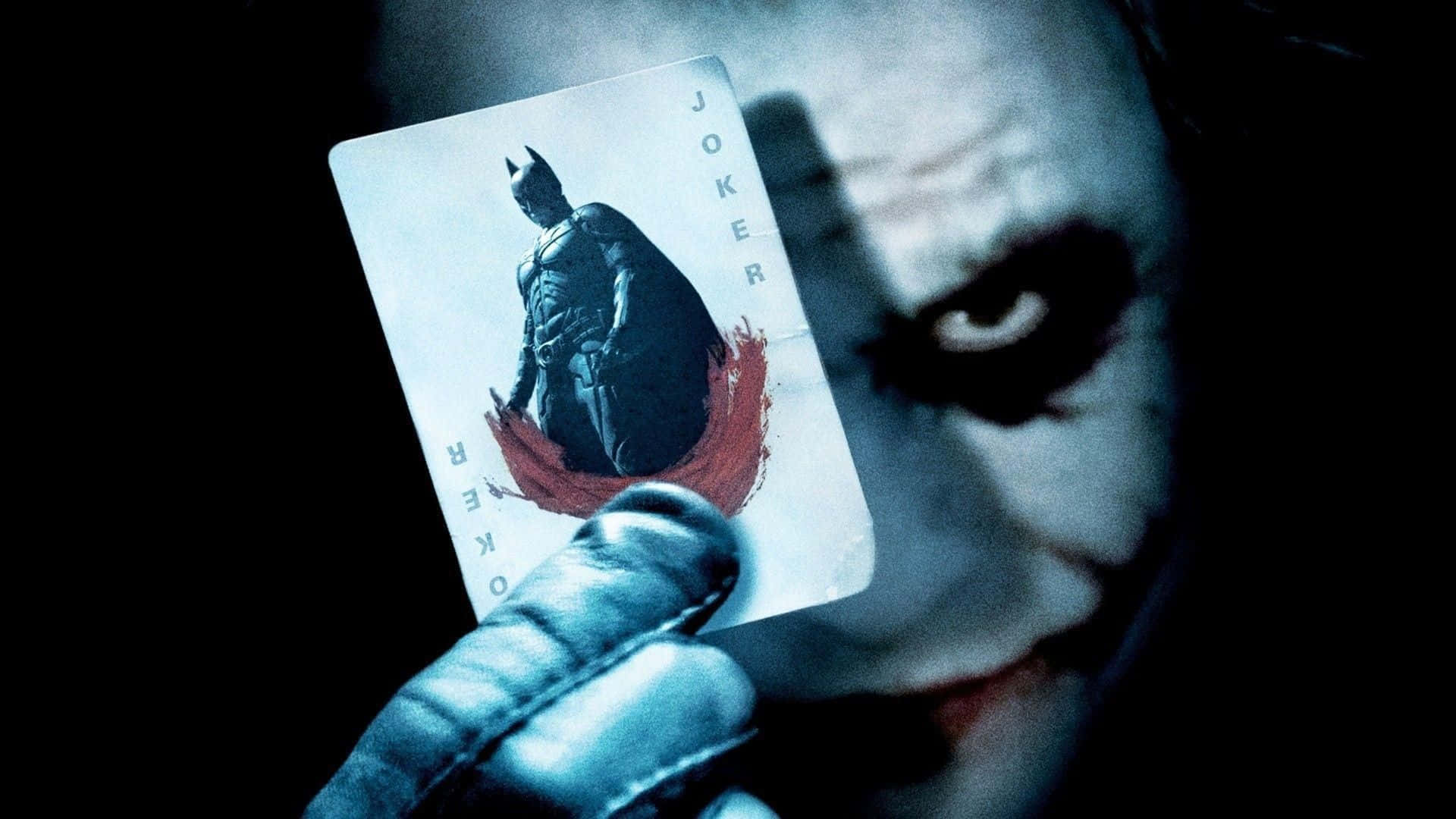 Joker Card 桌布 壁紙 — 一隻戴手套的手拿著一張印有蝙蝠俠嘅小丑啤牌，背景係黑暗，仲有小丑面孔嘅威脅性特寫。