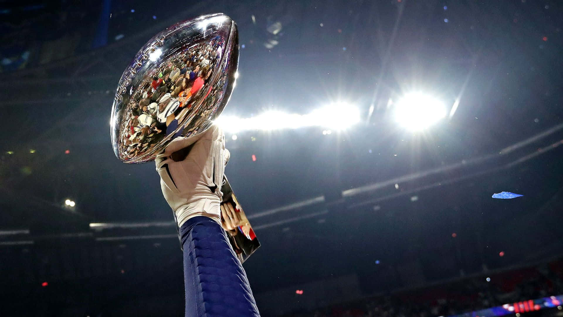 Super Bowl 桌布 壁紙 — 一隻戴著手套的手在明亮的體育館燈光下，伴隨著飄落的五彩紙屑，勝利地舉起閃閃發光的超級碗獎盃。