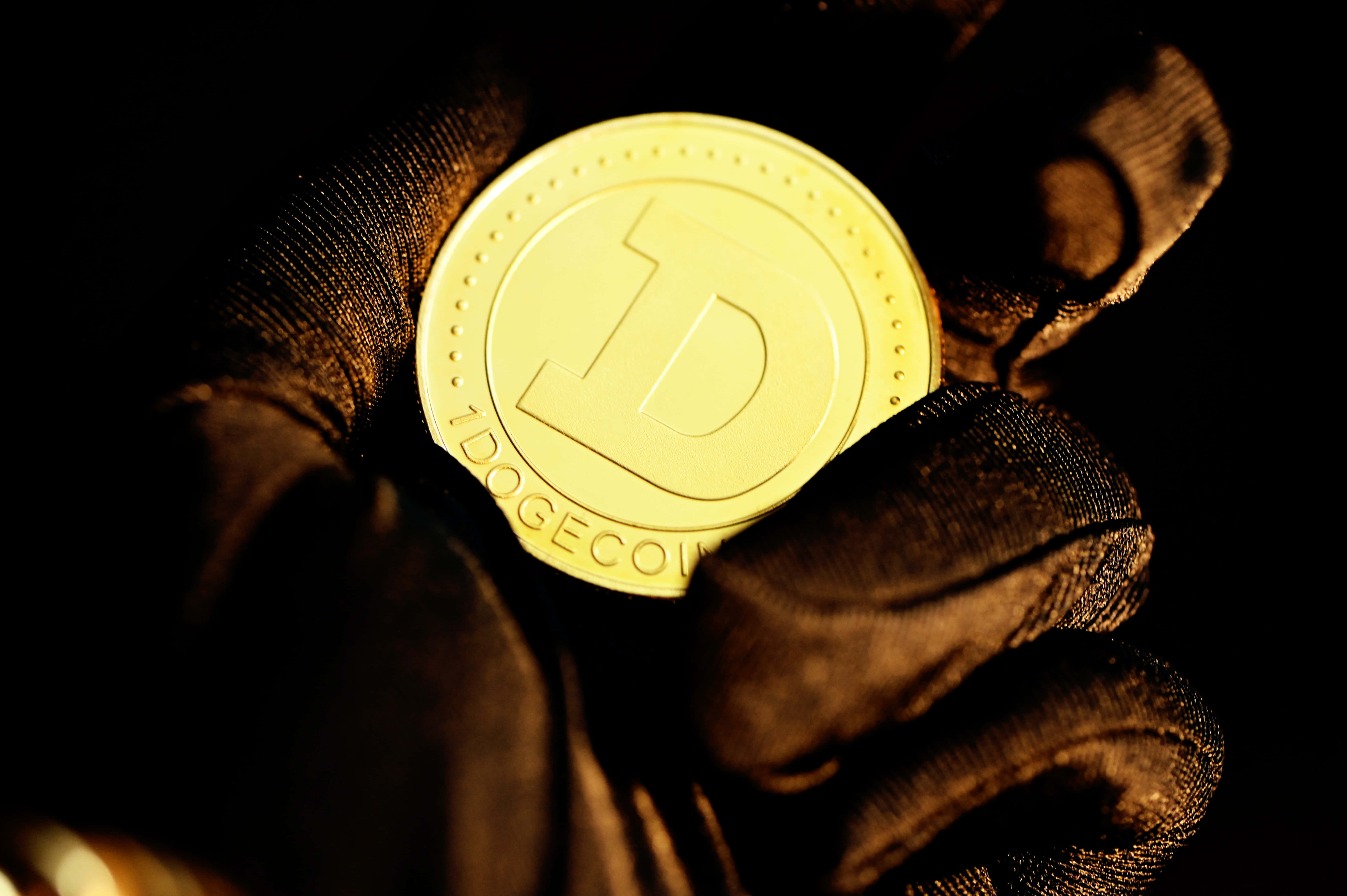 Dogecoin 桌布 壁紙 — 一隻戴黑色手套的手在黑暗背景中握著閃閃發光的金色Dogecoin。