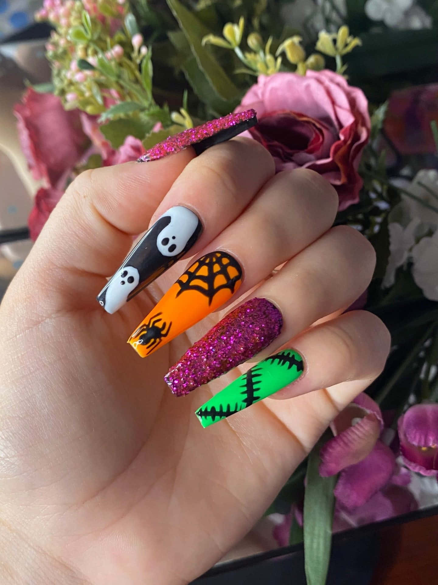 Halloween Nail Art 桌布 壁紙 — 一隻手，帶有充滿活力的萬聖節主題亞克力指甲，上面有鬼魂、蜘蛛、蜘蛛網和科學怪人設計，背景是粉紅玫瑰。