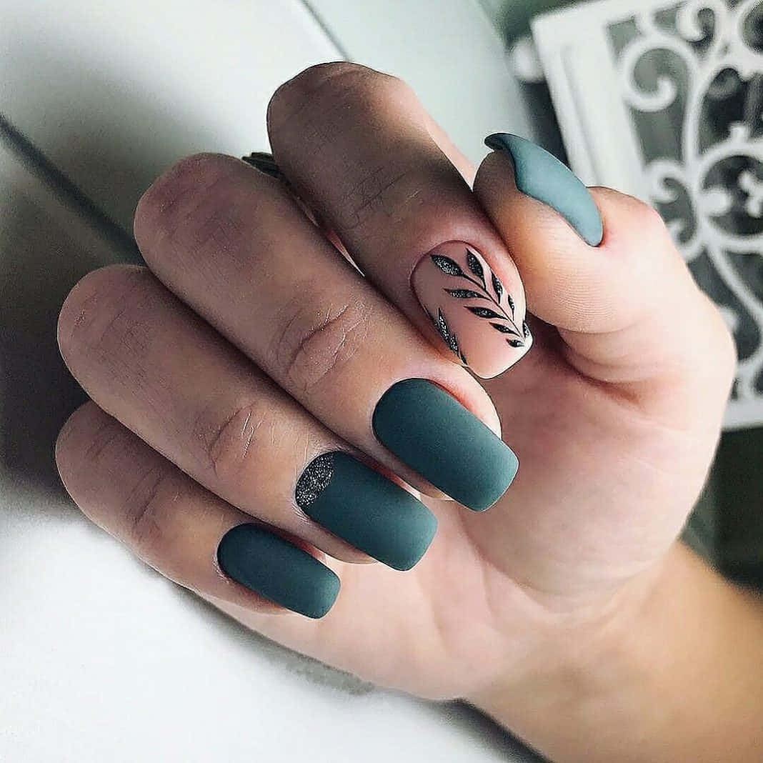 Spring Nails 桌布 壁紙 — 一隻手，帶有啞光青綠色指甲，飾有精緻的黑色葉子圖案和閃粉點綴，置於柔軟的白色表面上。