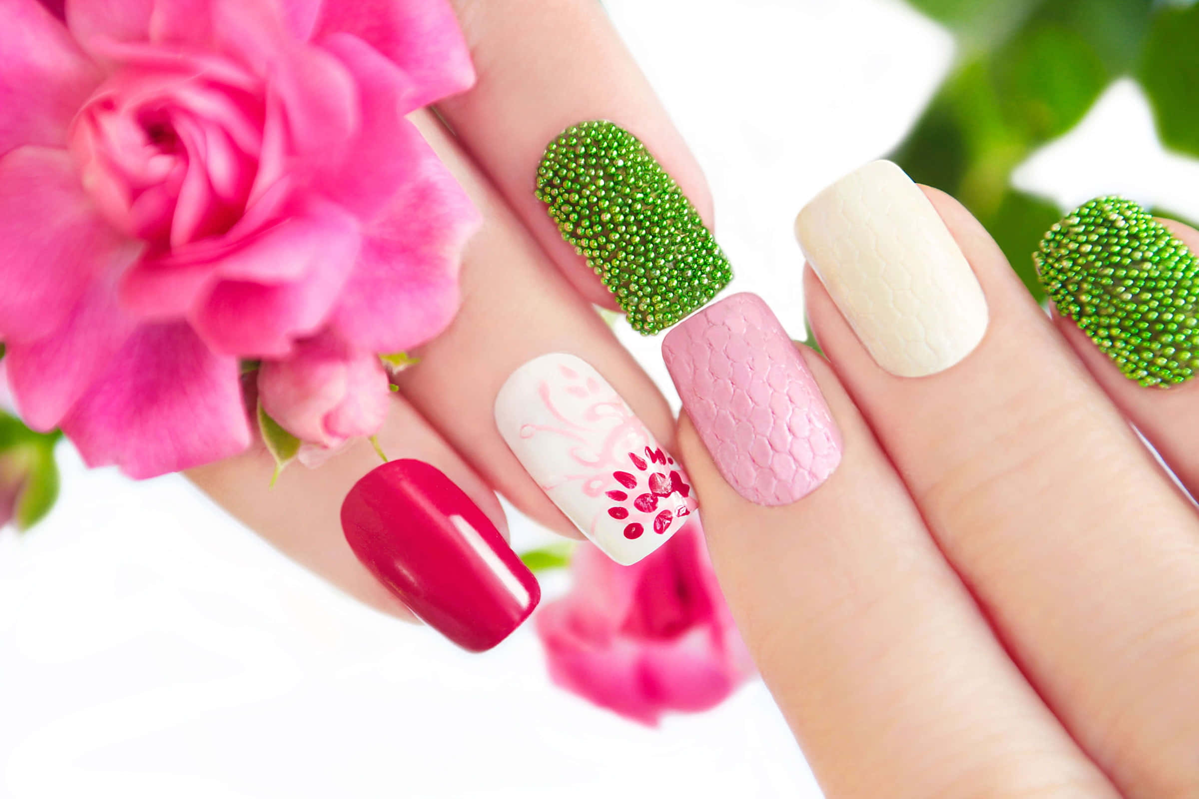 Cute Nails 桌布 壁紙 — 一隻手，帶有色彩繽紛的美甲，包括綠色珠子、粉紅色鱗片和花卉圖案，旁邊是一朵鮮豔的粉紅色玫瑰。