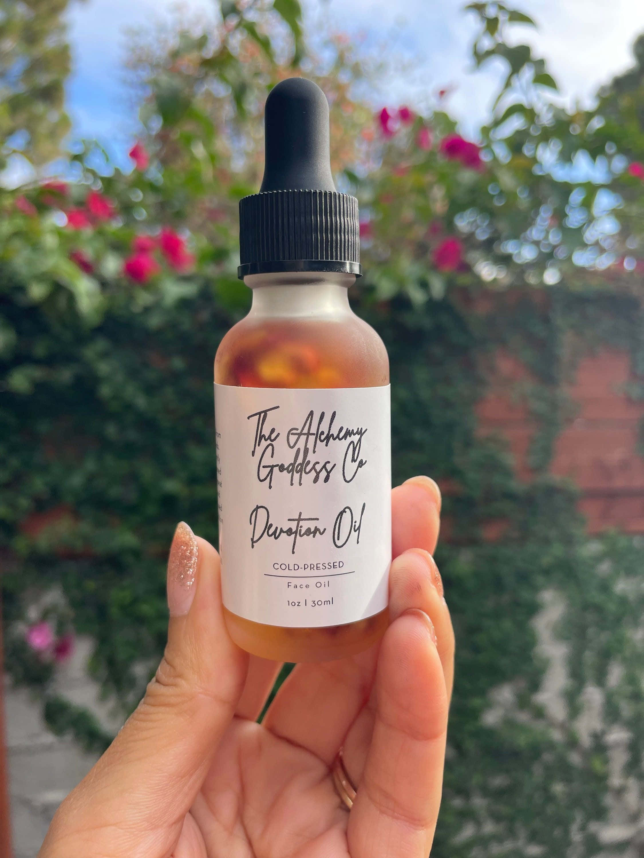 Rose Oil 桌布 壁紙 — 一隻手喺明亮日光下，以綠色樹葉同粉紅色花做背景，揸住一個標籤為「Devotion Oil」嘅琥珀色面部護理油樽。