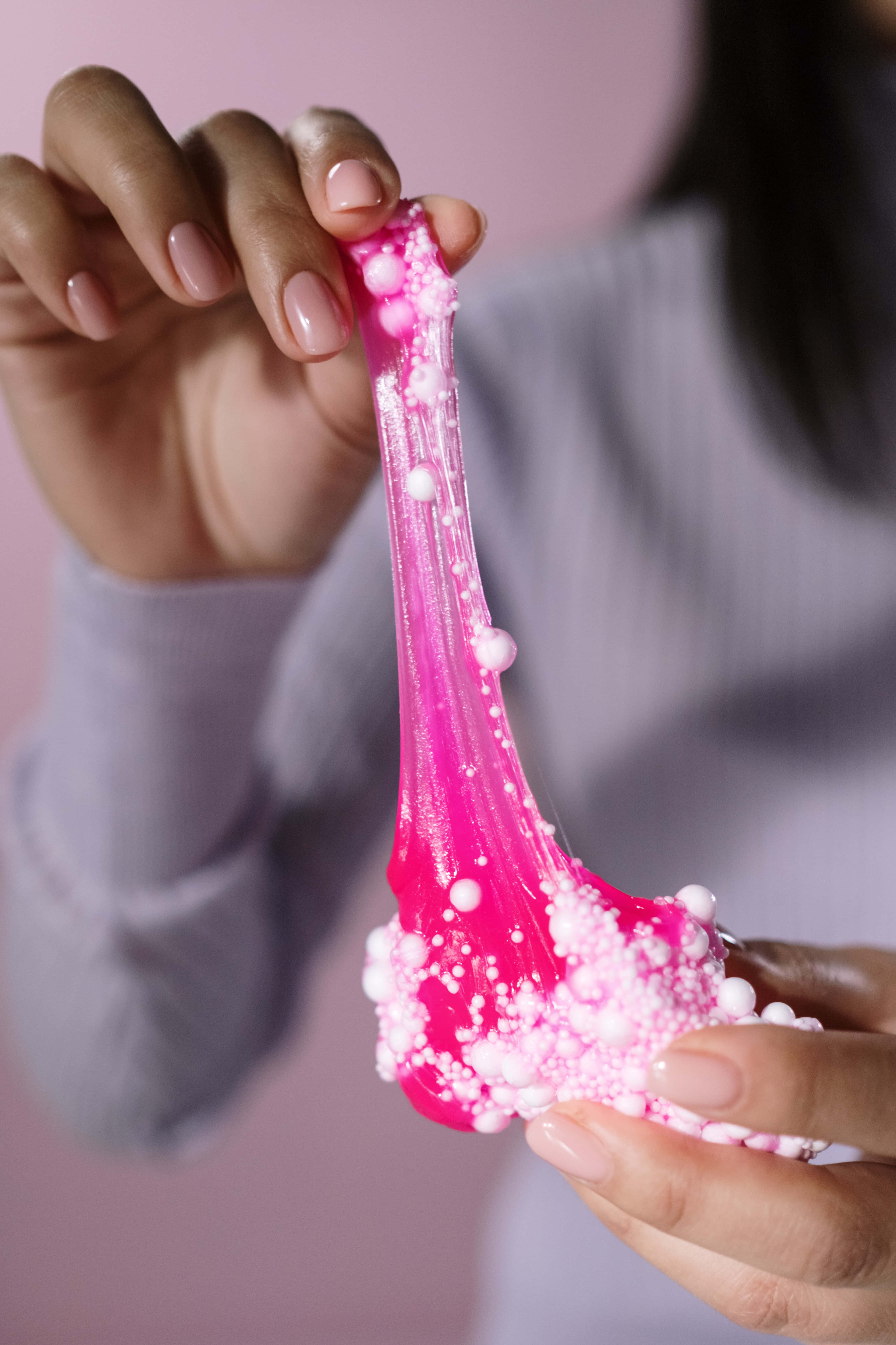 slime 桌布 壁紙 — 一隻手喺柔和粉紅色背景上拉伸充滿活力嘅粉紅色閃粉史萊姆，上面點綴住白色珠子。