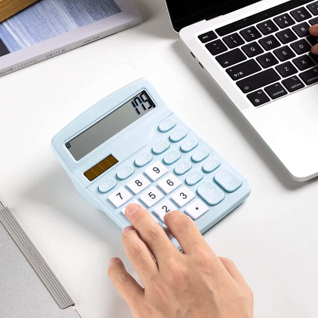 Calculator 桌布 壁紙 — 一隻手在乾淨的白色桌子上，按壓淺藍色計算機上的按鈕，旁邊有手提電腦和筆記本。