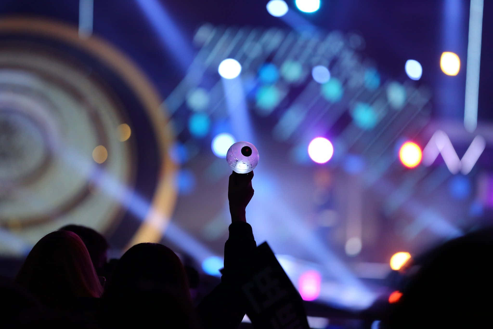 Bts Army Bomb 桌布 壁紙 — 一隻手在充滿活力的演唱會中舉起一個發光的燈球，周圍有色彩繽紛的舞台燈光和模糊的觀眾剪影。