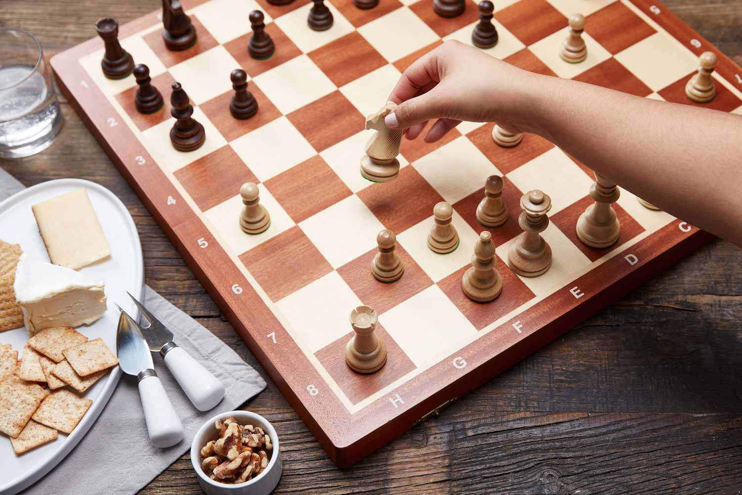 Chessboard 桌布 壁紙 — 一隻手在木製棋盤上移動國際象棋兵，旁邊放著一碟芝士餅乾和堅果，置於鄉村風格的桌子上。