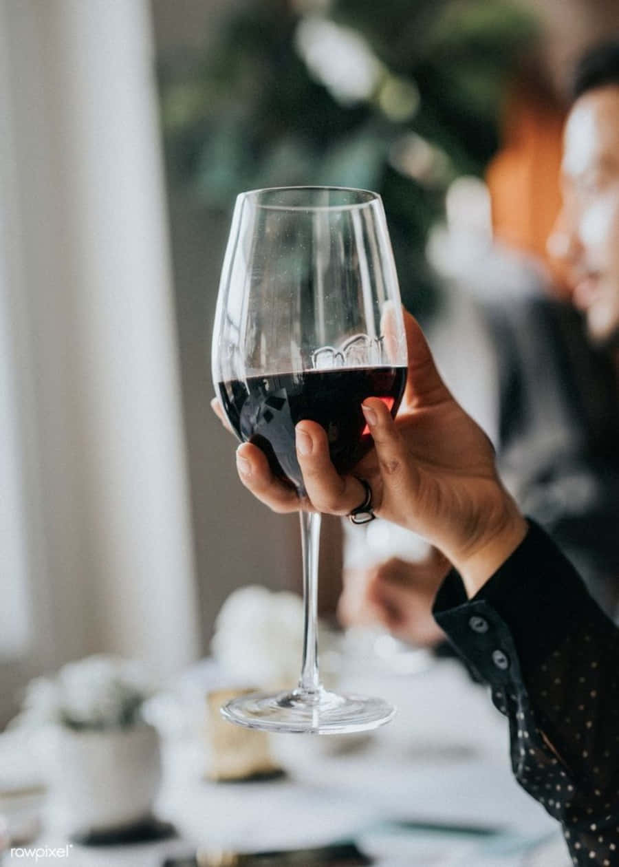 Black Wine 桌布 壁紙 — 一隻手在柔和光線的餐桌上拿著一杯深紅酒，背景有模糊的綠色植物和另一個人。