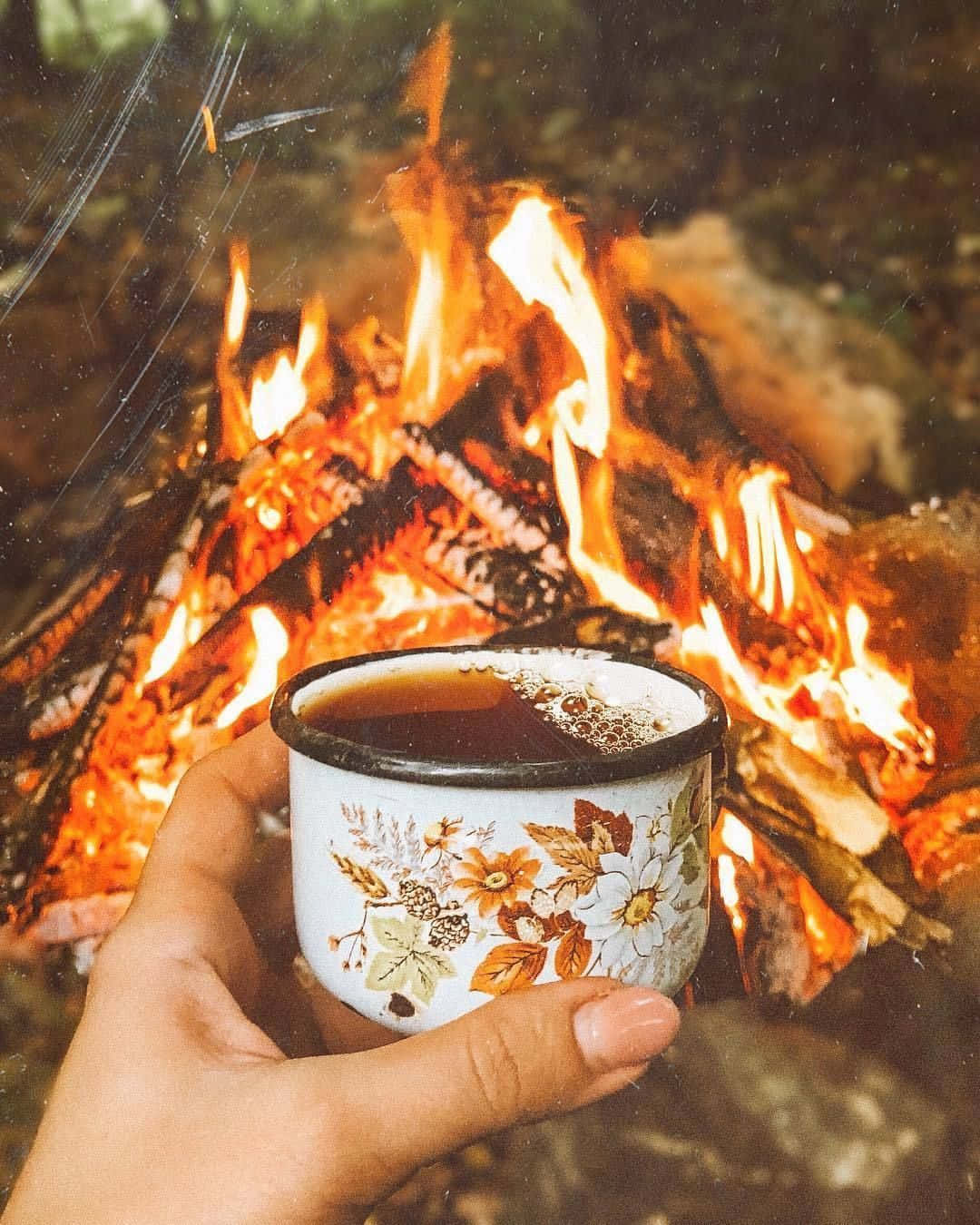 Fall Bonfire 桌布 壁紙 — 一隻手在森林中噼啪作響的營火前，拿著一個花卉圖案的琺瑯杯，裡面裝著熱飲。