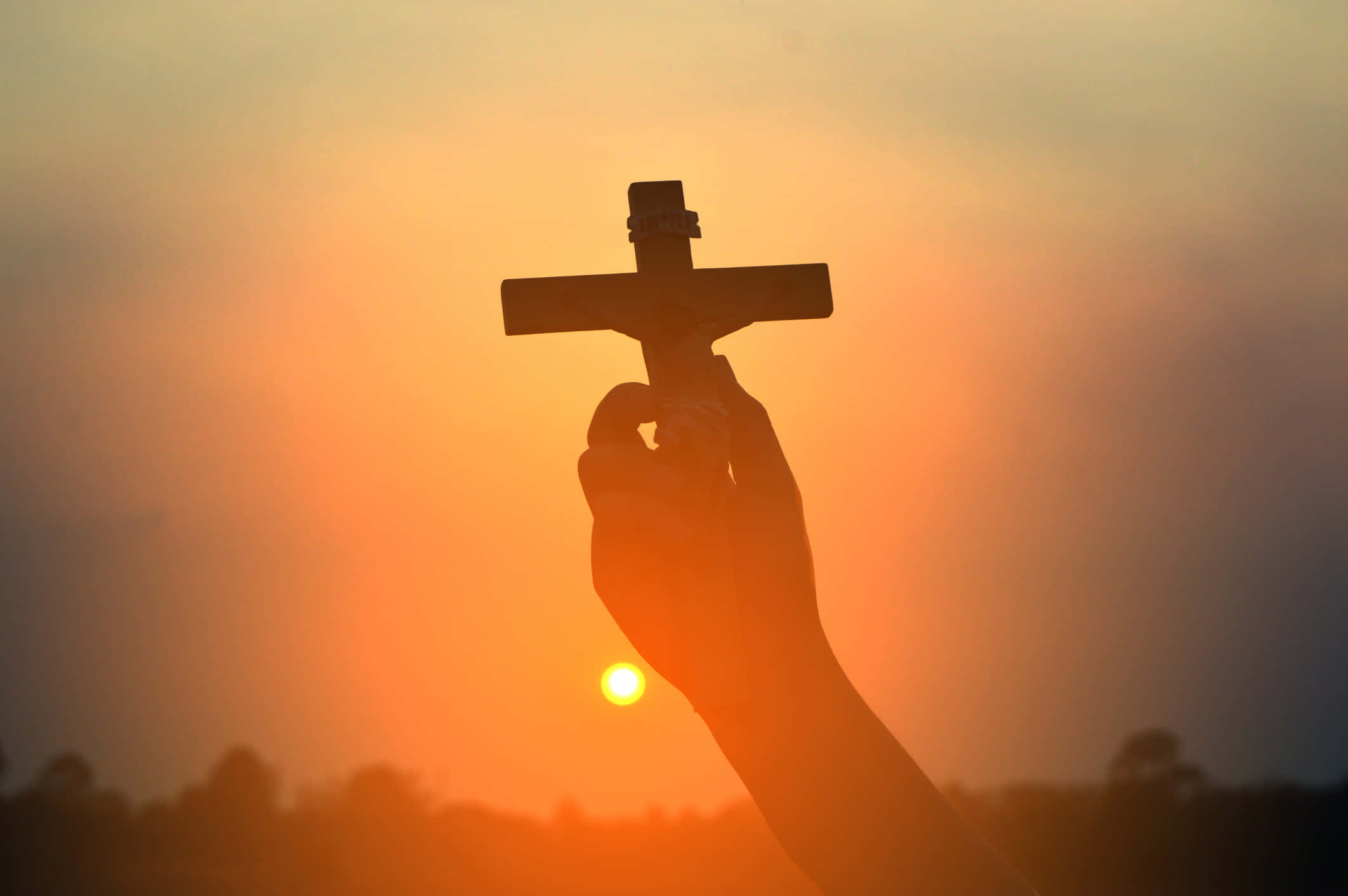 Religious Easter 桌布 壁紙 — 一隻手在溫暖的橙色和黃色光芒的日落天空下舉著十字架。