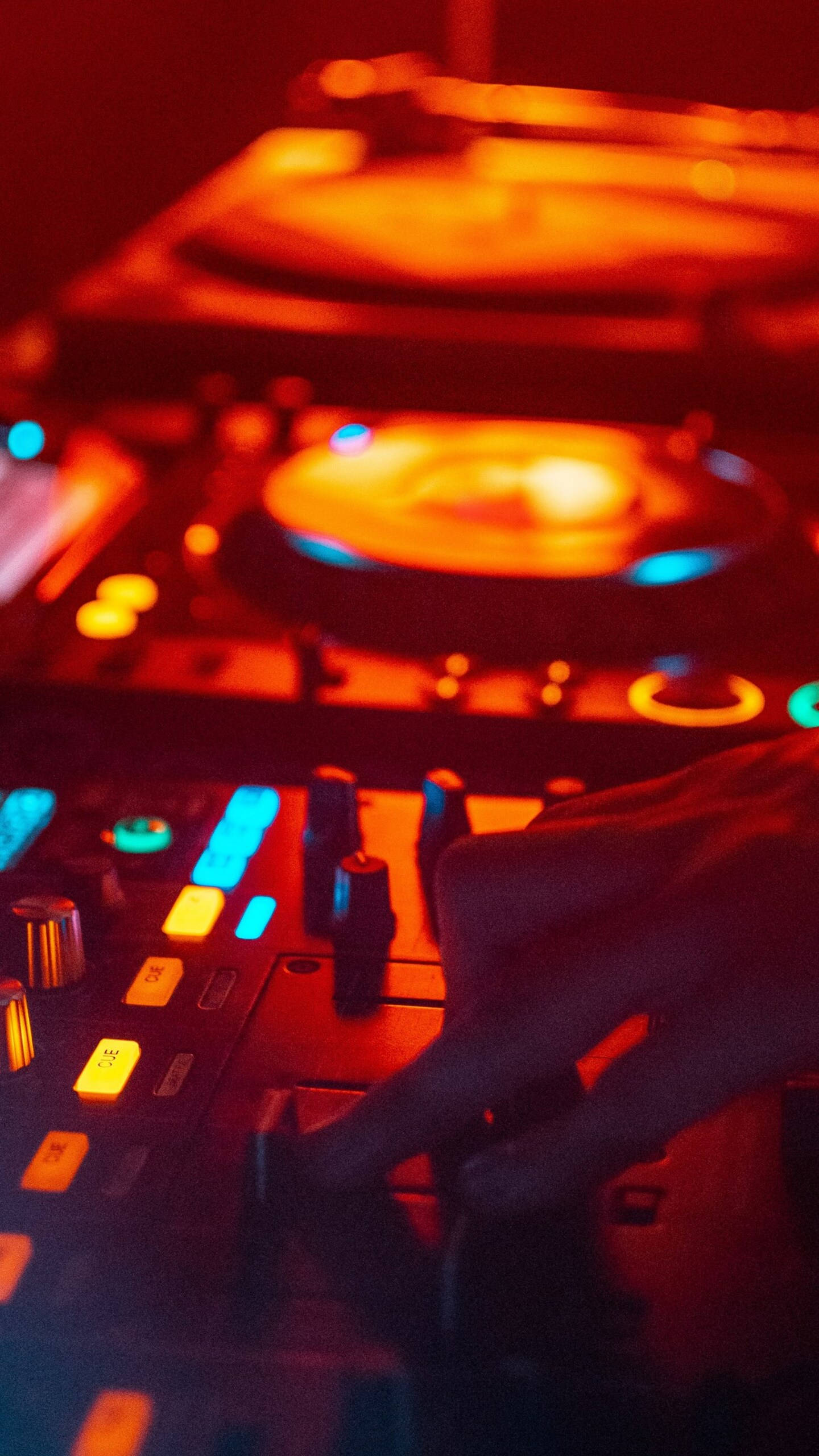 iPhone 12 Pro Max DJ 壁紙 — 一隻手在溫暖的紅色和冷藍色霓虹燈光下調整發光的DJ控制器上的旋鈕。