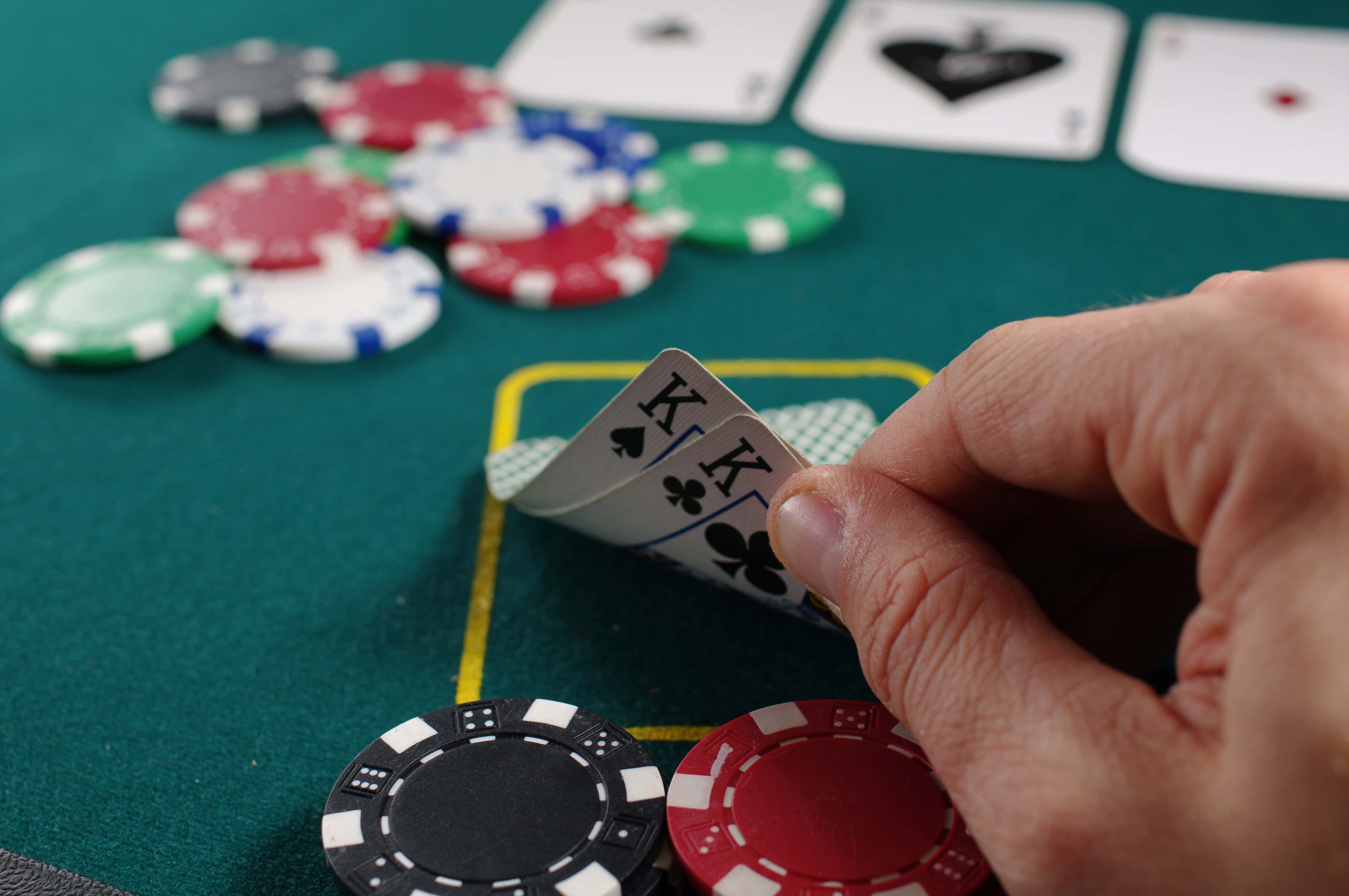 Poker Table 桌布 壁紙 — 一隻手在綠色嘅啤牌枱上，枱面散落住彩色籌碼同揭開嘅啤牌，手上揸住兩張梅花K。