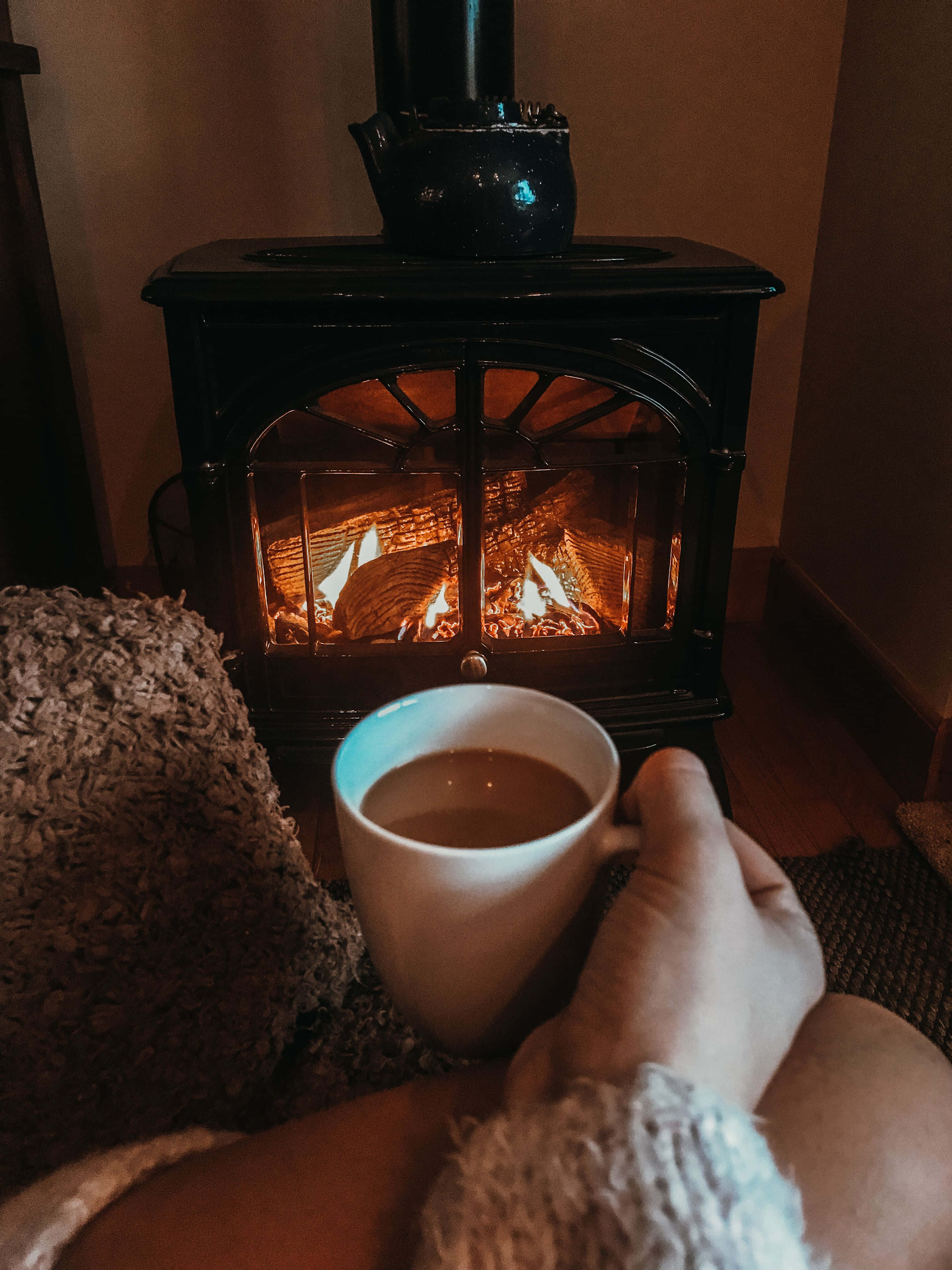 Cozy Fire 桌布 壁紙 — 一隻手在舒適昏暗的房間裡，捧著一杯溫暖的馬克杯，旁邊是發光的柴火爐。