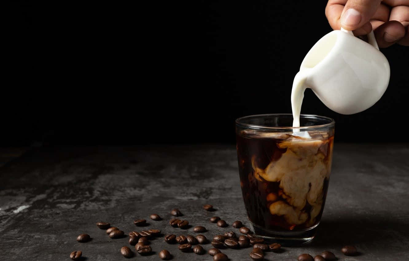 Milk And Coffee 桌布 壁紙 — 一隻手將奶油牛奶倒入一杯冰咖啡中，周圍散落著咖啡豆，置於深色紋理表面上。