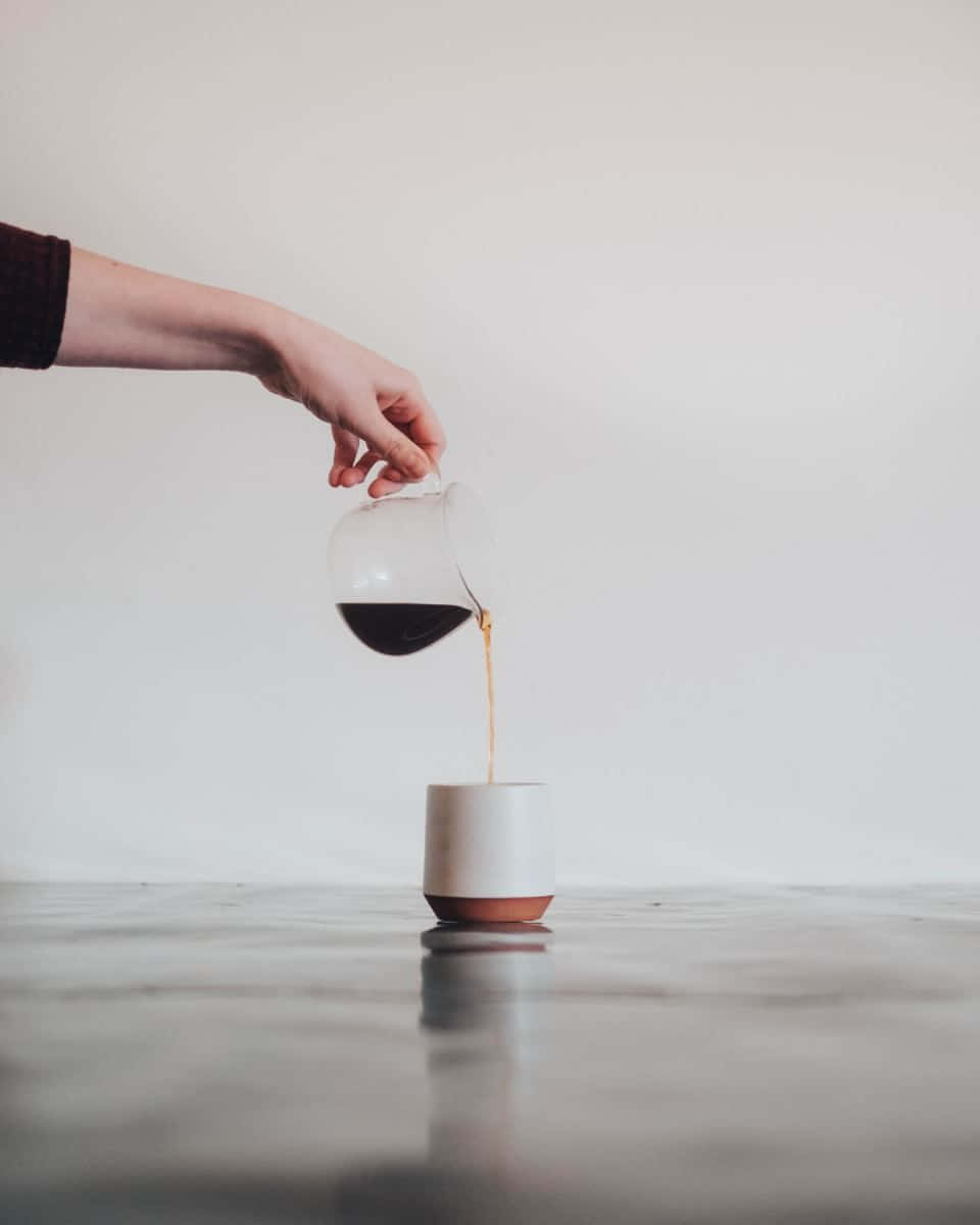 Minimalist Coffee 桌布 壁紙 — 一隻手將玻璃壺中的濃黑咖啡倒入簡約陶瓷杯中，背景為純白牆壁。