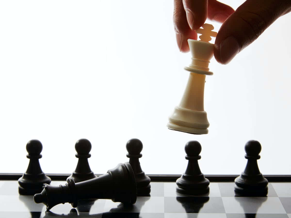 Black And White Chess 桌布 壁紙 — 一隻手將白色國王棋舉起，高於倒下的黑色國王和站立的兵卒，在棋盤上，背景明亮。