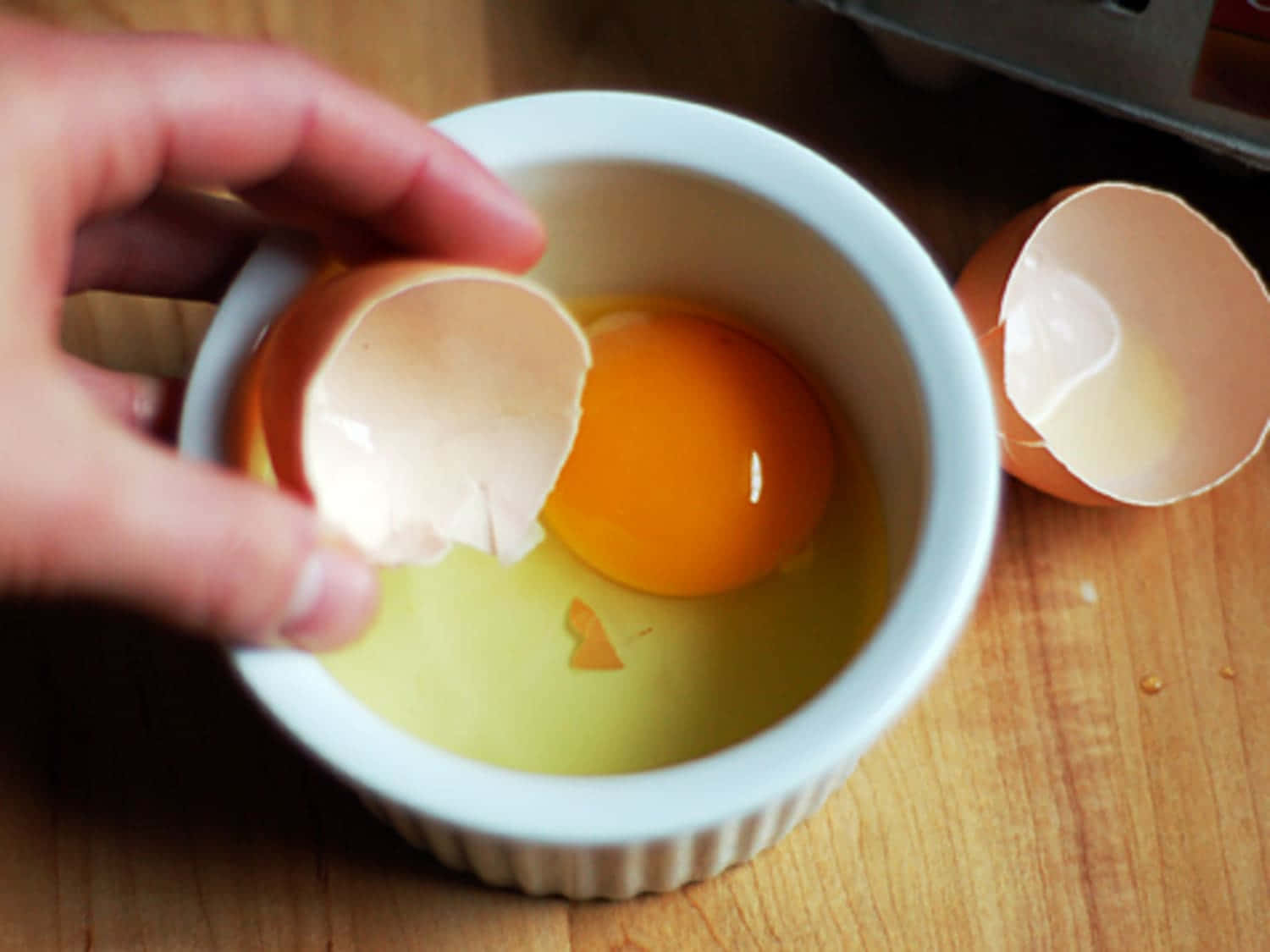 Eggshell 桌布 壁紙 — 一隻手小心地將蛋殼放入一個白色的小烤碗中，碗內有鮮豔的橙色蛋黃和清澈的蛋白，放在木質表面上。