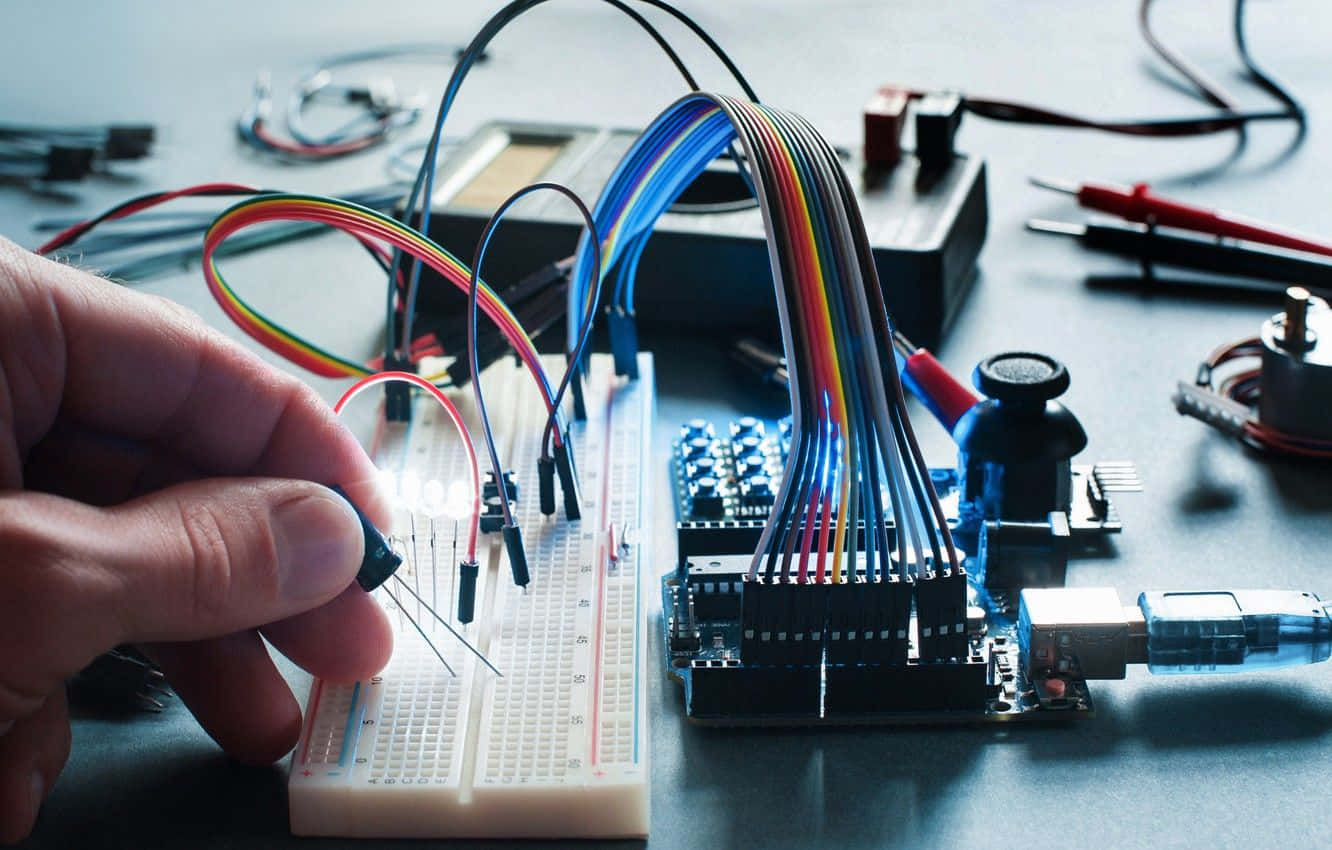 Electronics 桌布 壁紙 — 一隻手小心翼翼地將發光的LED連接到麵包板上，周圍纏繞著五顏六色的電線和電子元件。