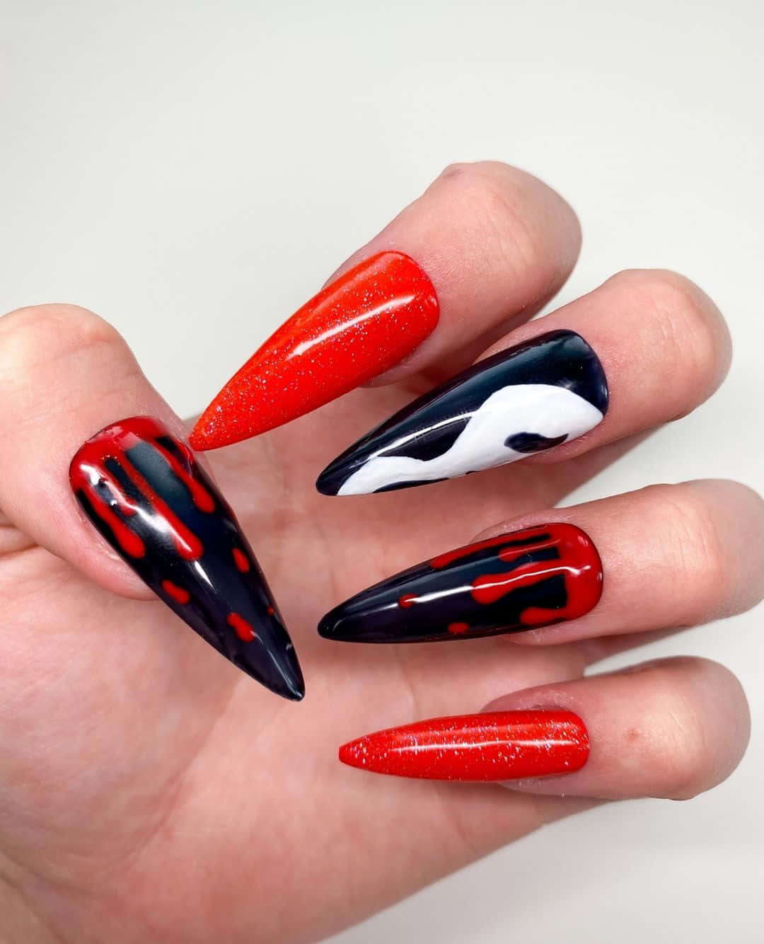 Halloween Nail Art 桌布 壁紙 — 一隻手展示大膽的細高跟指甲，帶有紅色閃粉、黑色和白色抽象設計，背景為純白色。