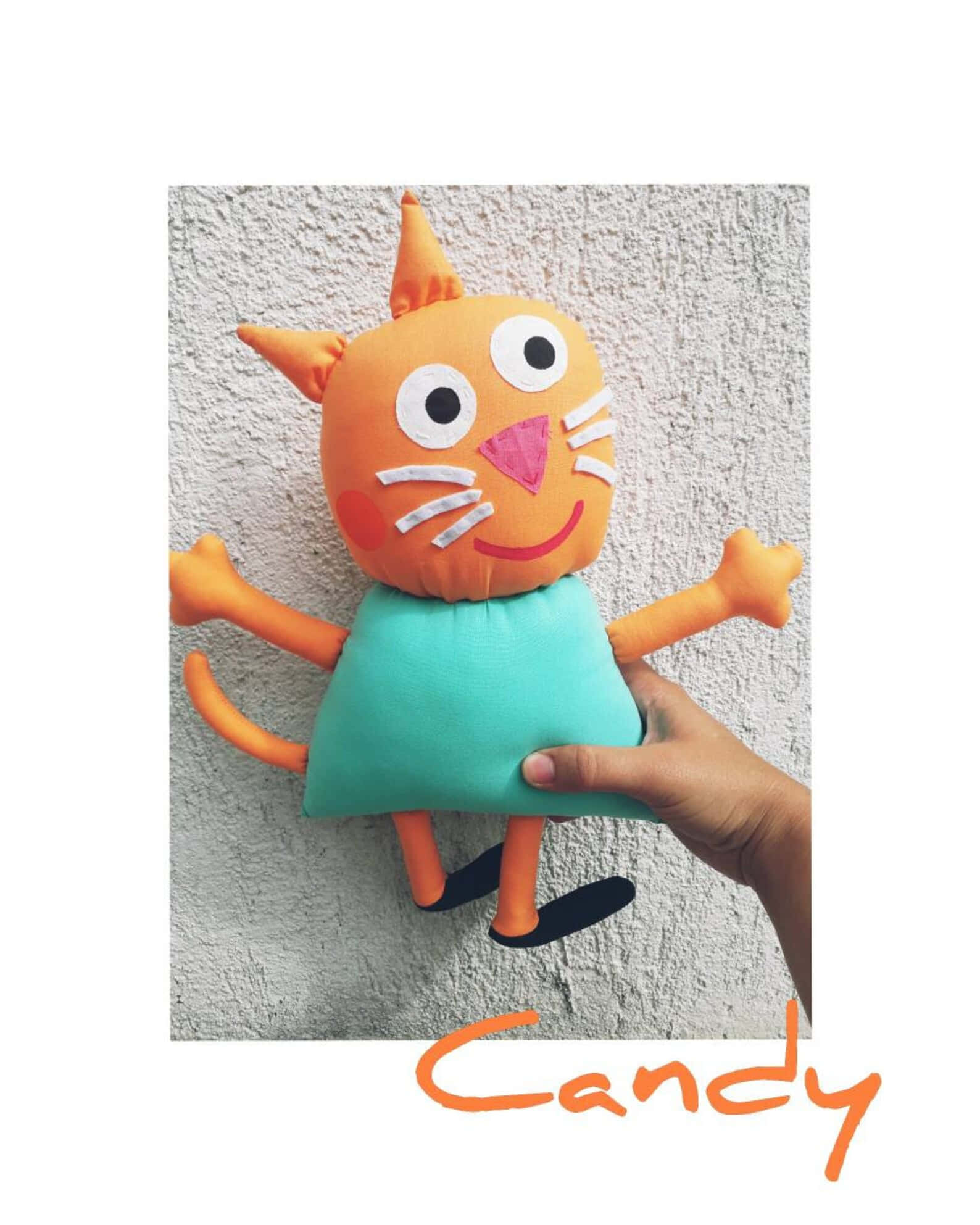 Candy Cat 桌布 壁紙 — 一隻手工製作的橙色貓咪毛公仔穿著青綠色連衣裙，被舉起靠在有紋理的灰色牆上。