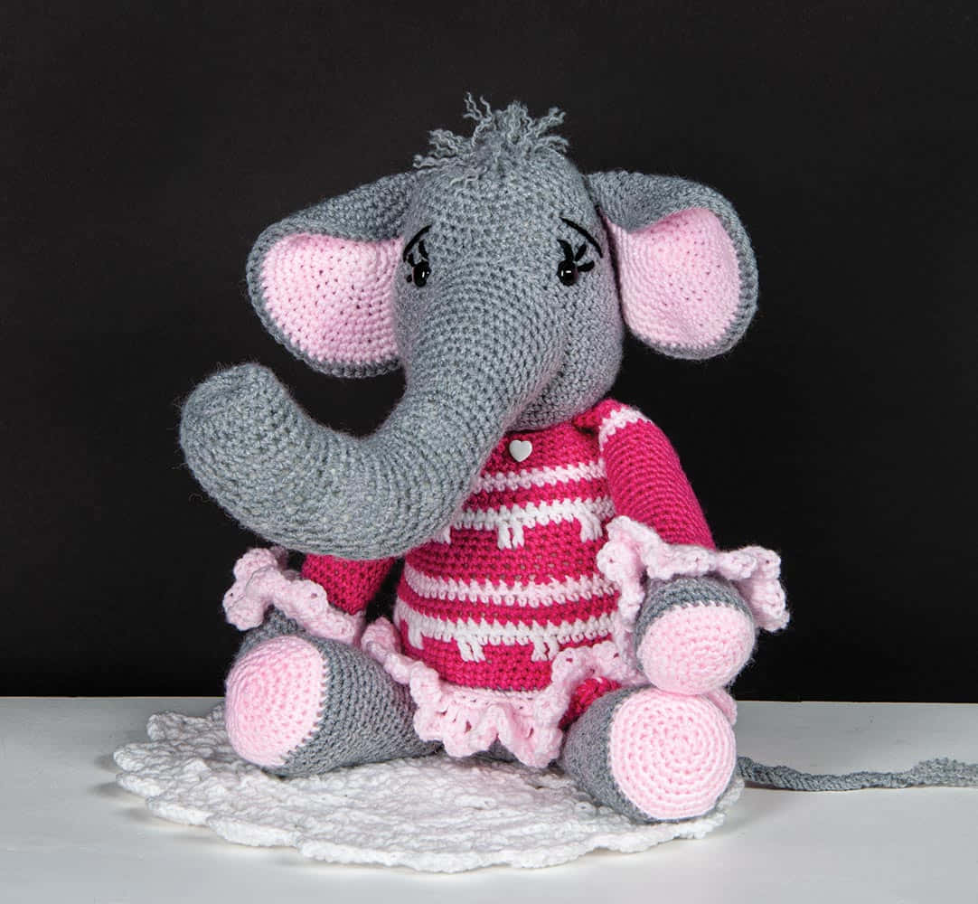 Emily Elephant 桌布 壁紙 — 一隻手工鉤織的大象，穿著粉紅色條紋連衣裙，坐在黑色背景上的白色鏤空墊上。
