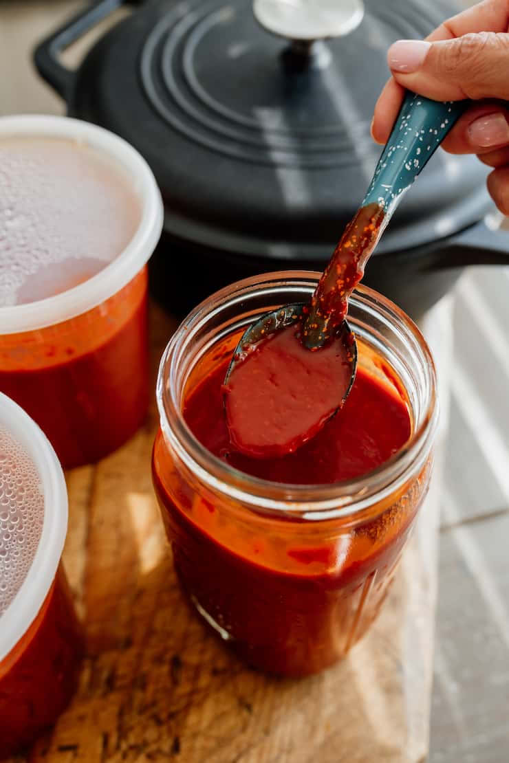 Red Sauce 桌布 壁紙 — 一隻手從木板上的一個玻璃罐中舀起一匙濃稠的紅色醬汁，附近還有其他容器。