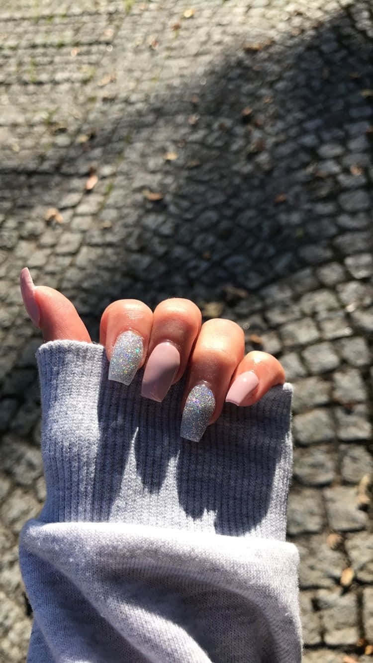 Cute Nails 桌布 壁紙 — 一隻手從灰色袖子中伸出，指甲帶有閃亮銀色和啞光紫紅色，背景是陽光普照的鵝卵石小徑。