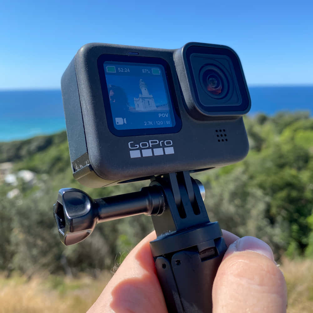 gopro 桌布 壁紙 — 一隻手拿著GoPro相機，在清澈藍天下捕捉燈塔俯瞰碧綠海洋和綠色山丘的景色。