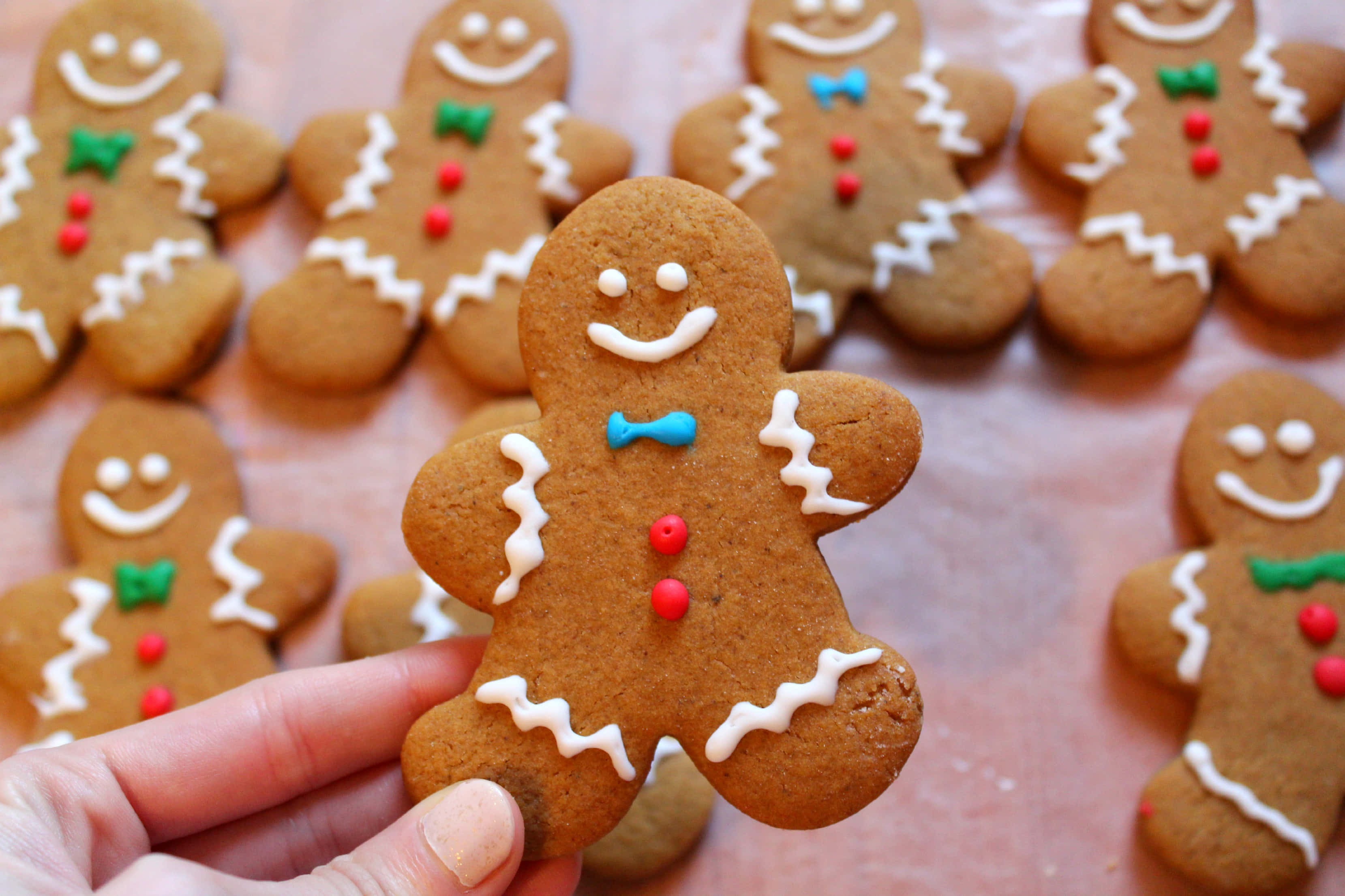 Gingerbread 桌布 壁紙 — 一隻手拿著一個微笑的薑餅人曲奇，上面裝飾著白色糖霜和彩色糖果，背景是類似的曲奇。