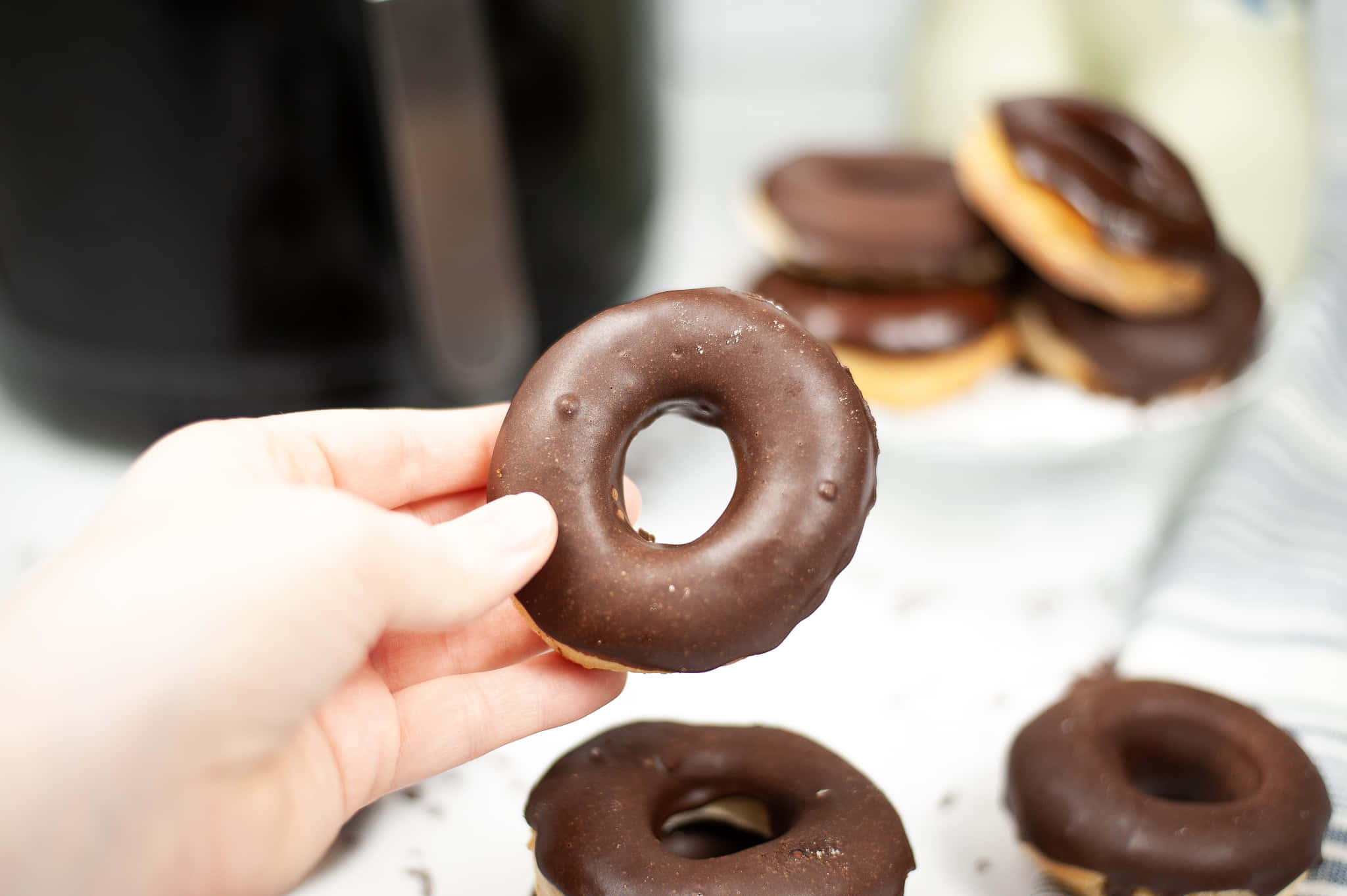 Glazed Donut 桌布 壁紙 — 一隻手拿著一個有光澤的朱古力釉面冬甩，其他冬甩散落在附近的白色表面上。