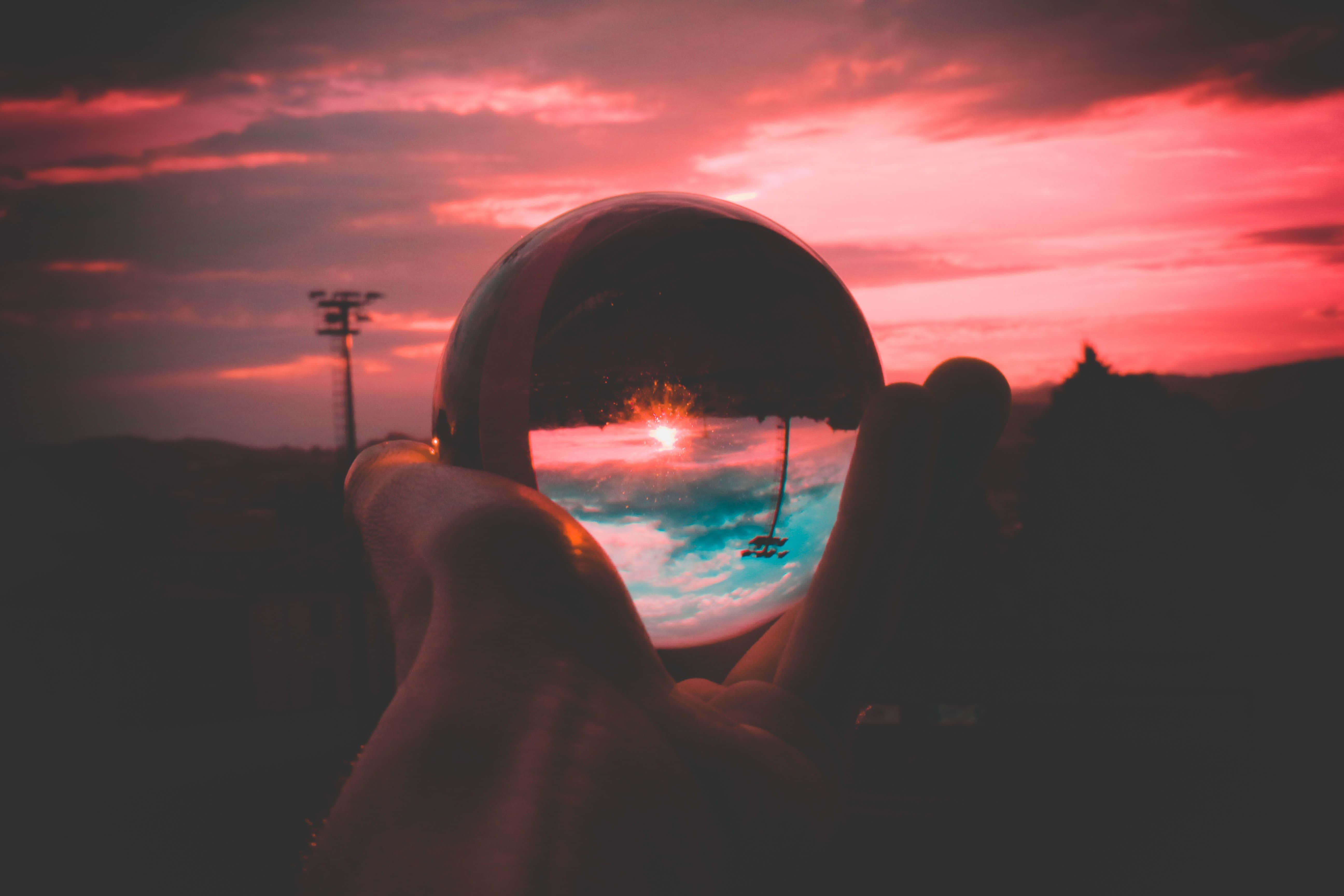 Crystal Ball 桌布 壁紙 — 一隻手拿著一個水晶球，反射出水面上 vibrant 的日落和帆船，背景中有一座電線塔。