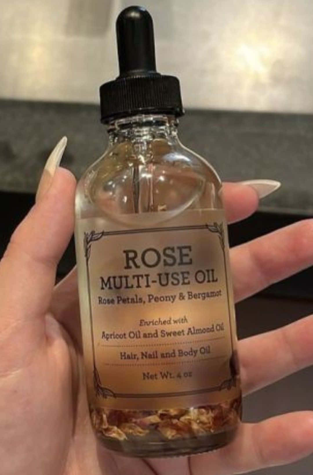 Rose Oil 桌布 壁紙 — 一隻手拿著一個玫瑰多用途油玻璃瓶，底部有乾花瓣和黑色滴管蓋。