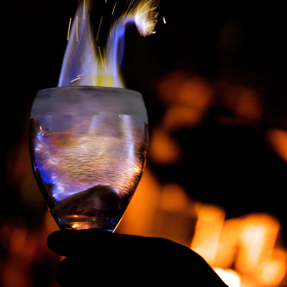 Beautiful Drink 桌布 壁紙 — 一隻手拿著一個玻璃杯，杯緣噴出發光的藍色火焰，背景是溫暖模糊的火焰。