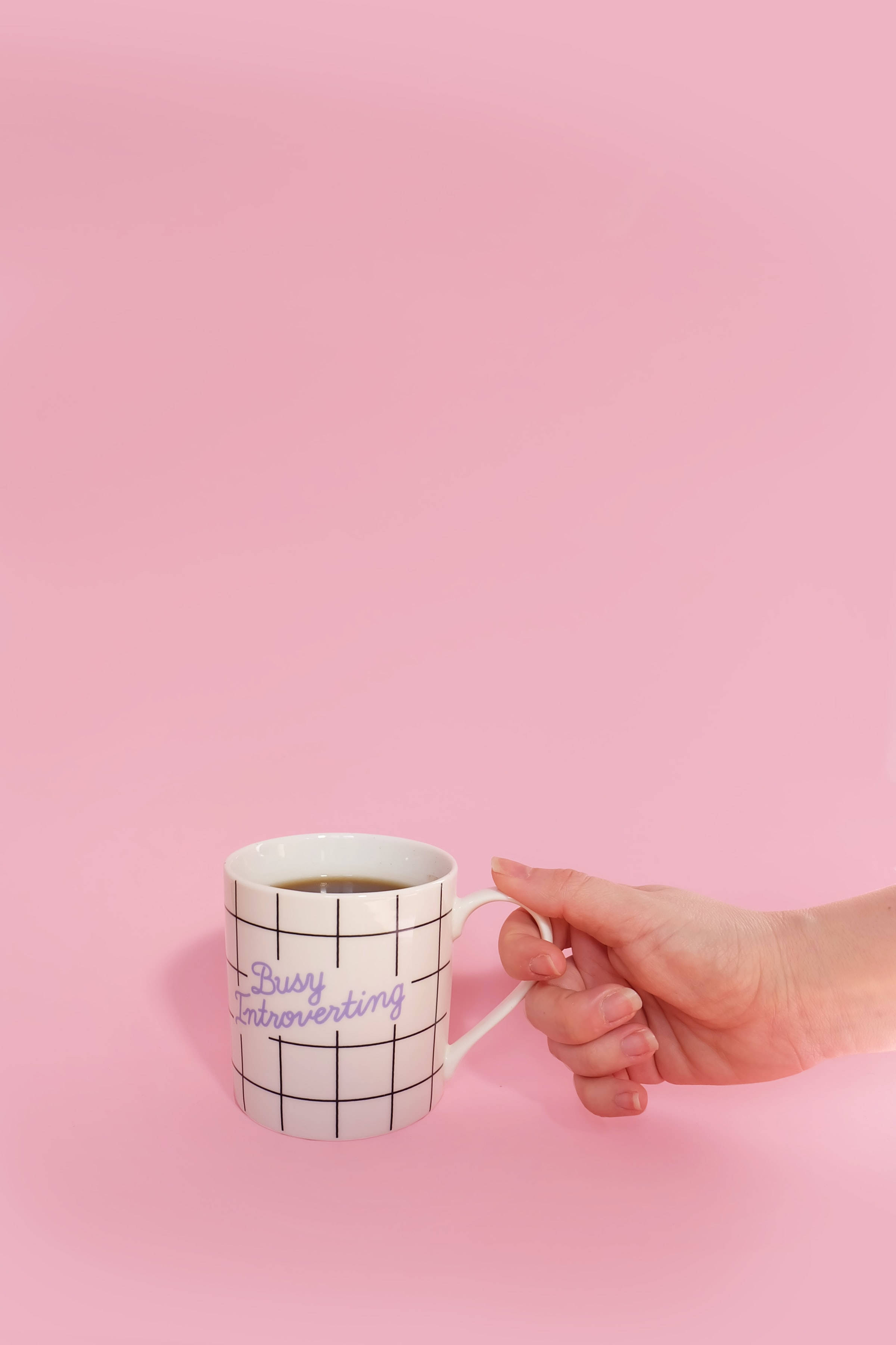 mug 桌布 壁紙 — 一隻手拿著一個白色馬克杯，上面有黑色格紋和紫色「Busy Introverting」文字，背景為純粉色。