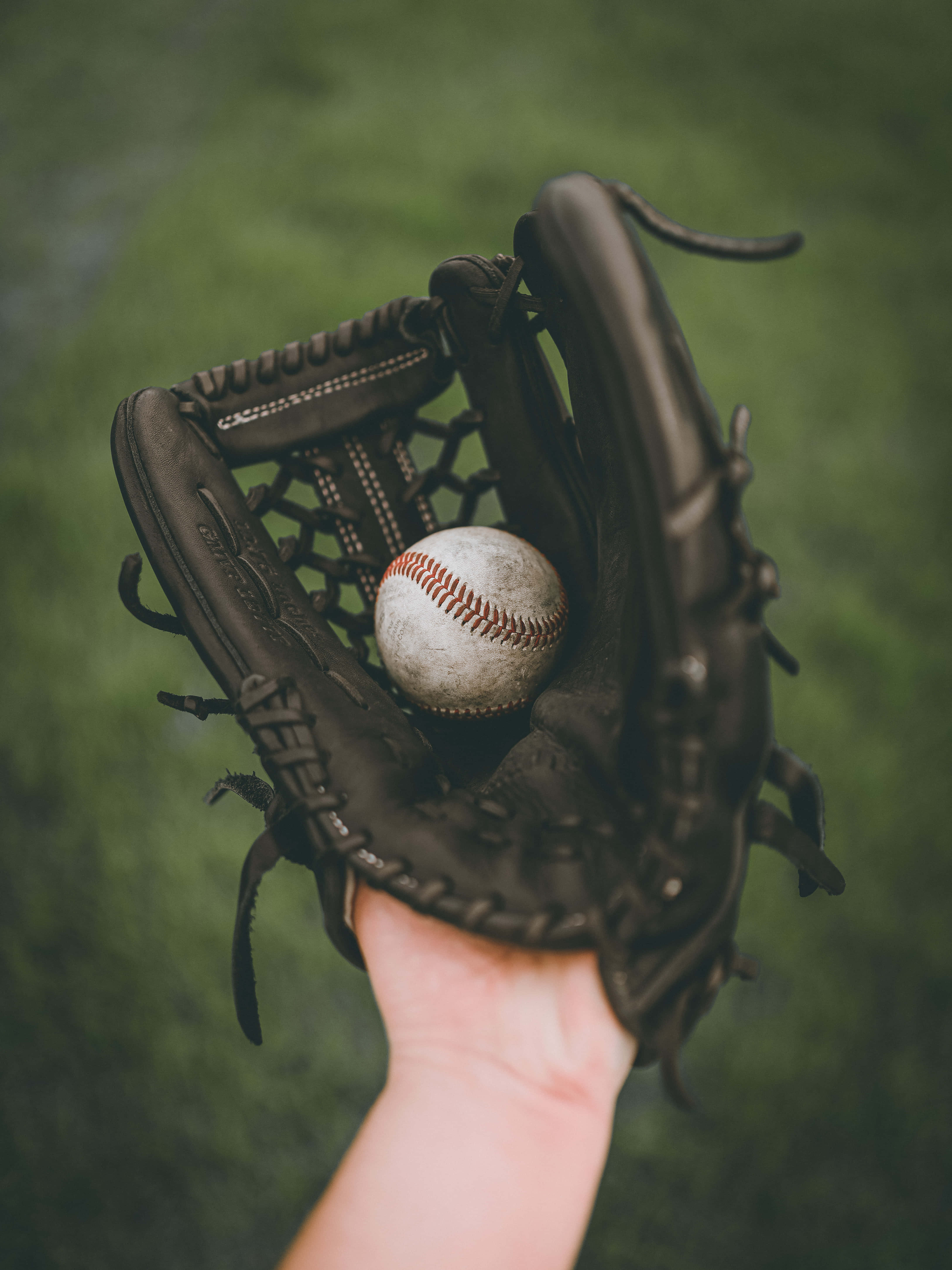 Baseball Gloves 桌布 壁紙 — 一隻手拿著一個破舊的黑色棒球手套，懷裡抱著一個用得很舊、帶紅色縫線的白色棒球，背景是模糊的綠色球場。