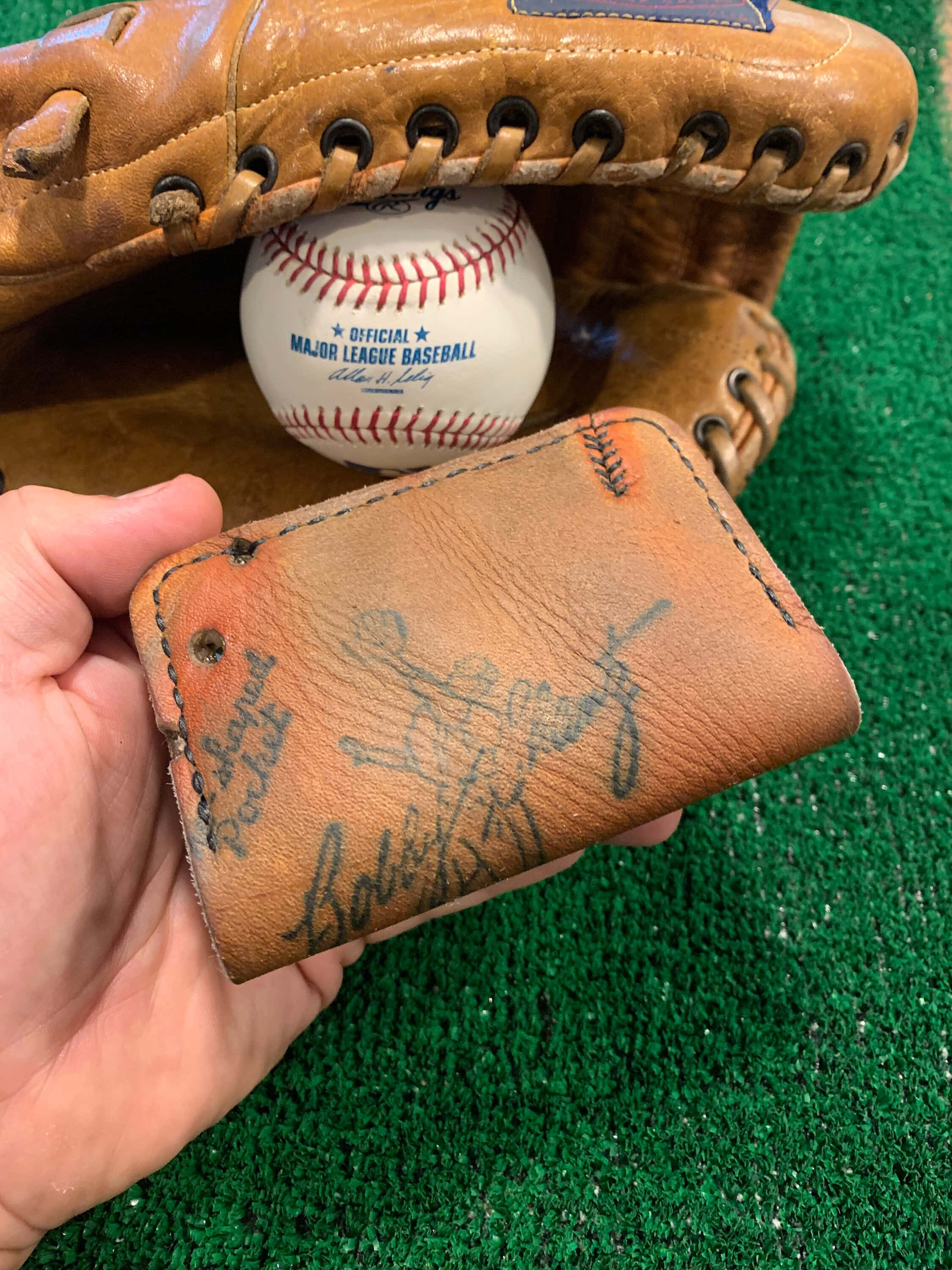 Baseball Gloves 桌布 壁紙 — 一隻手拿著一個磨損的皮革錢包，上面有褪色的簽名，背景是綠色草地，附近有一個棒球和手套。