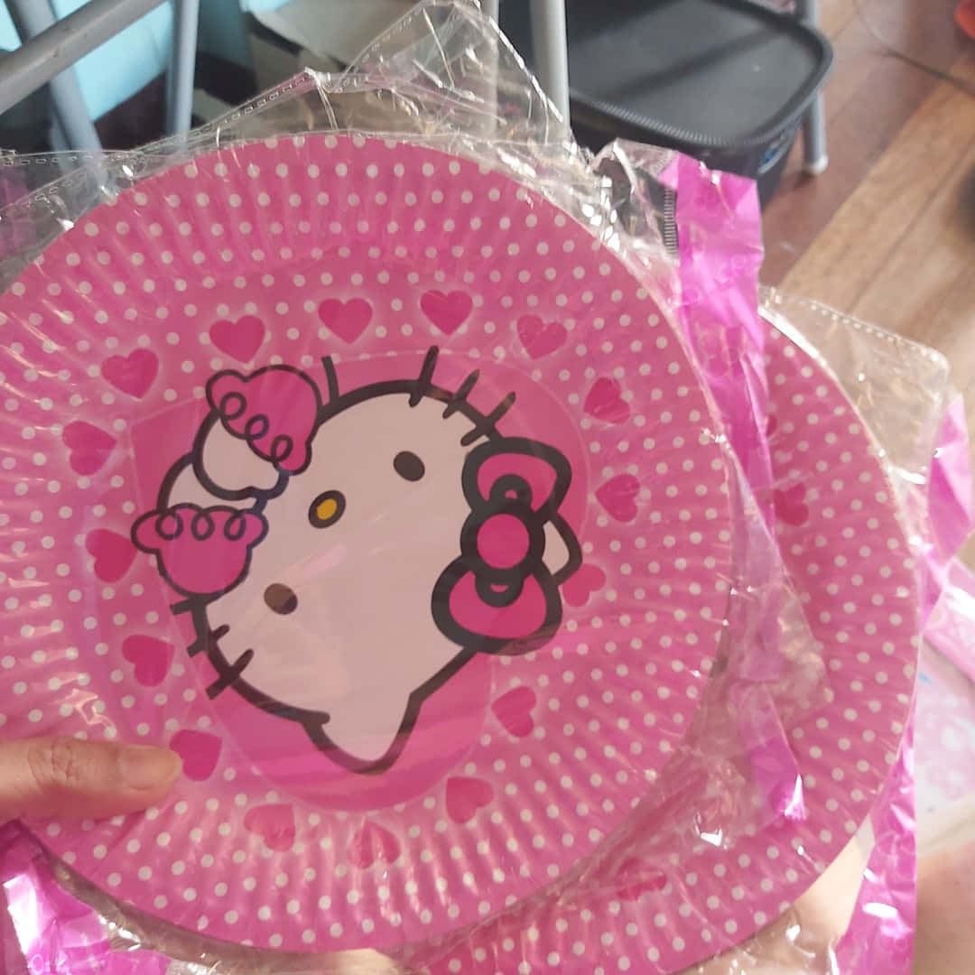 Hello Kitty Party 桌布 壁紙 — 一隻手拿著一個粉紅色Hello Kitty紙碟，上面有白色圓點和心形圖案，並用透明塑膠包裝著。