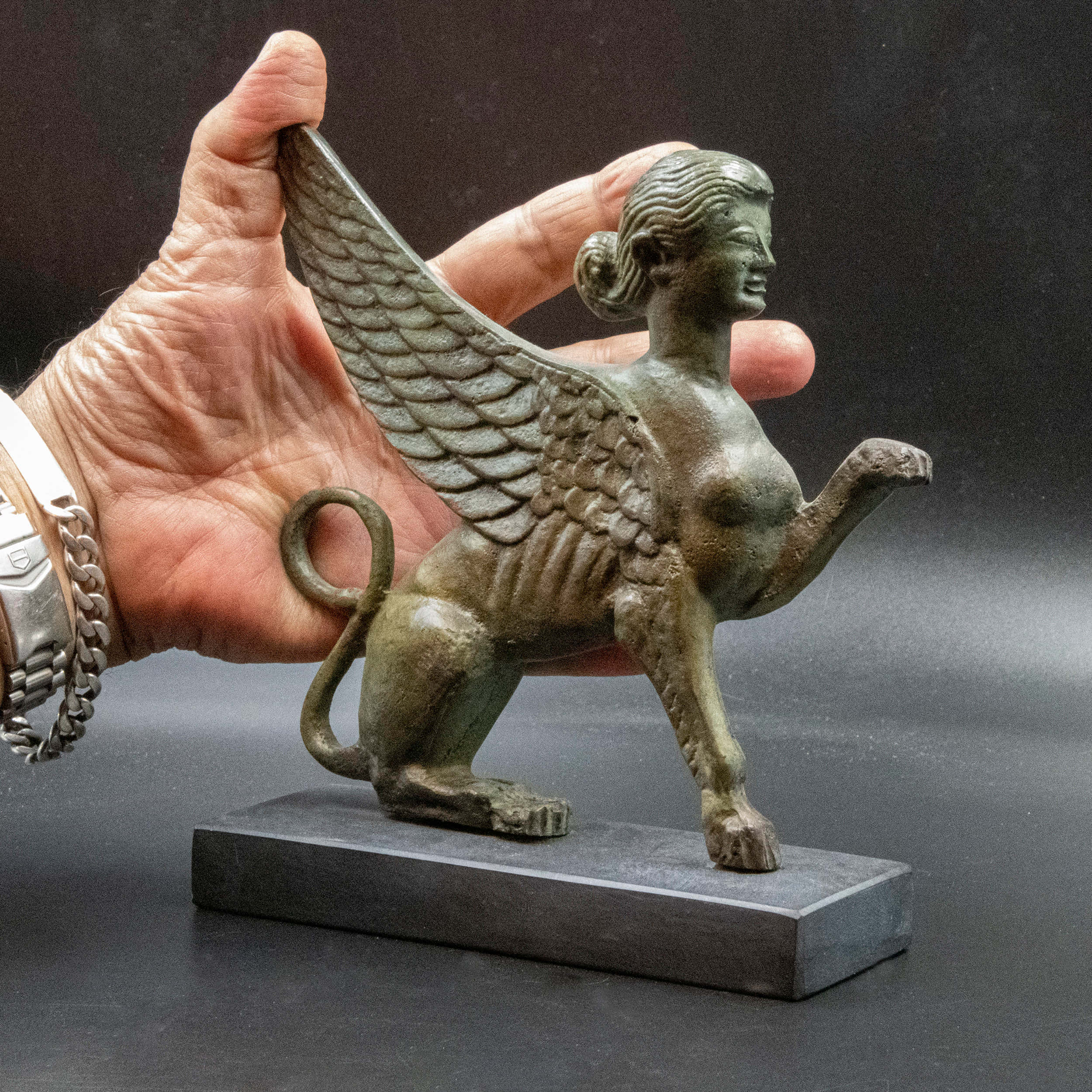 Mystical Artifacts 桌布 壁紙 — 一隻手拿著一個細節豐富的青銅獅身人面像，帶有翅膀和獅身，背景為深色工作室。