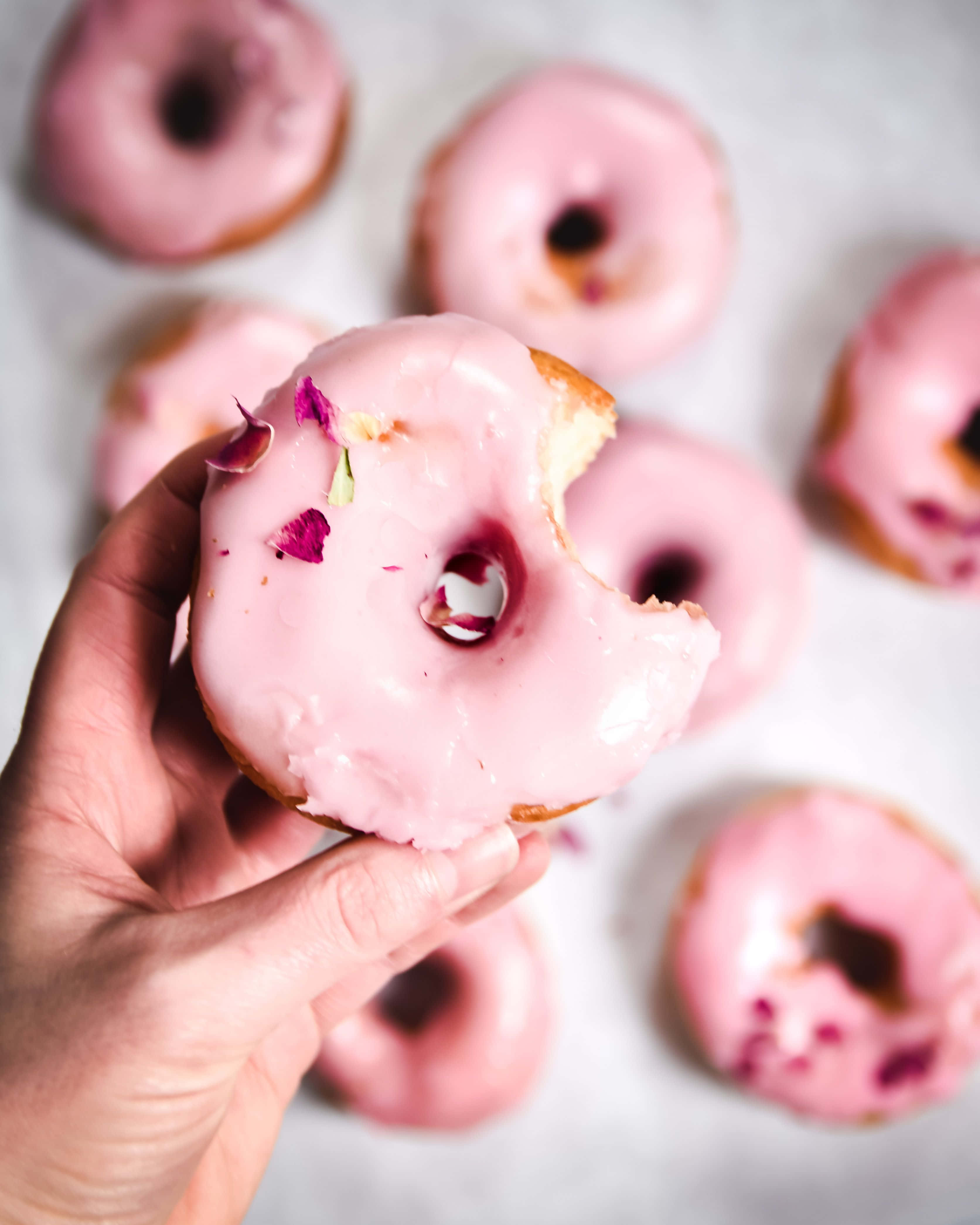 Glazed Donut 桌布 壁紙 — 一隻手拿著一個被咬過的粉紅色糖霜甜甜圈，上面撒有玫瑰花瓣，背景是模糊的類似甜甜圈。